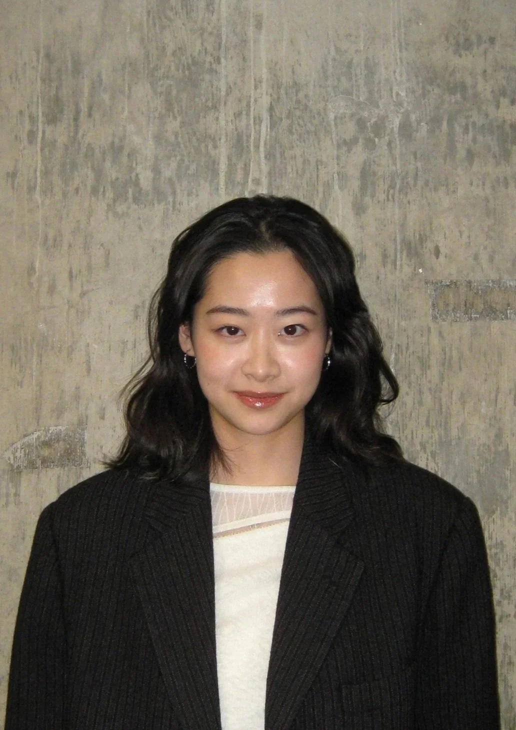 Portrait of a person with medium-length dark hair, wearing a black blazer over a white top, standing against a textured wall.