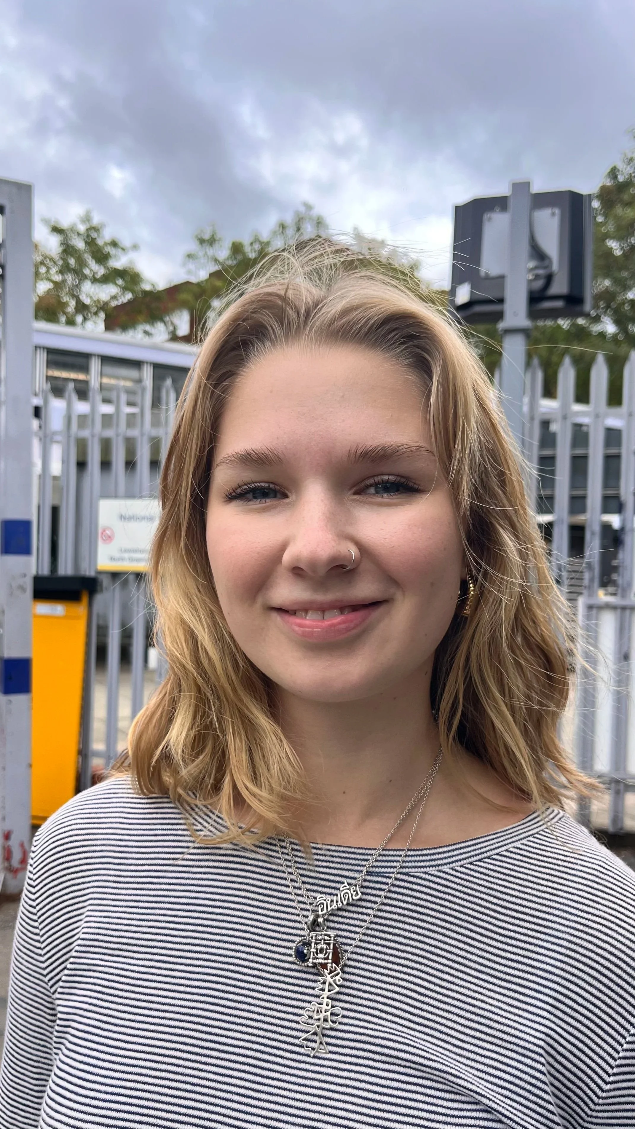 Portrait of as person with shoulder-length blonde hair, wearing a striped shirt, standing outdoors in front of a metal fence and a cloudy sky.