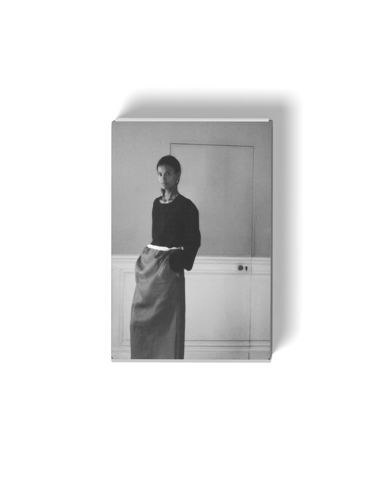 Black and white photograph of a woman standing indoors, looking at the camera, with hands in her pockets, wearing a dark top and a long skirt.