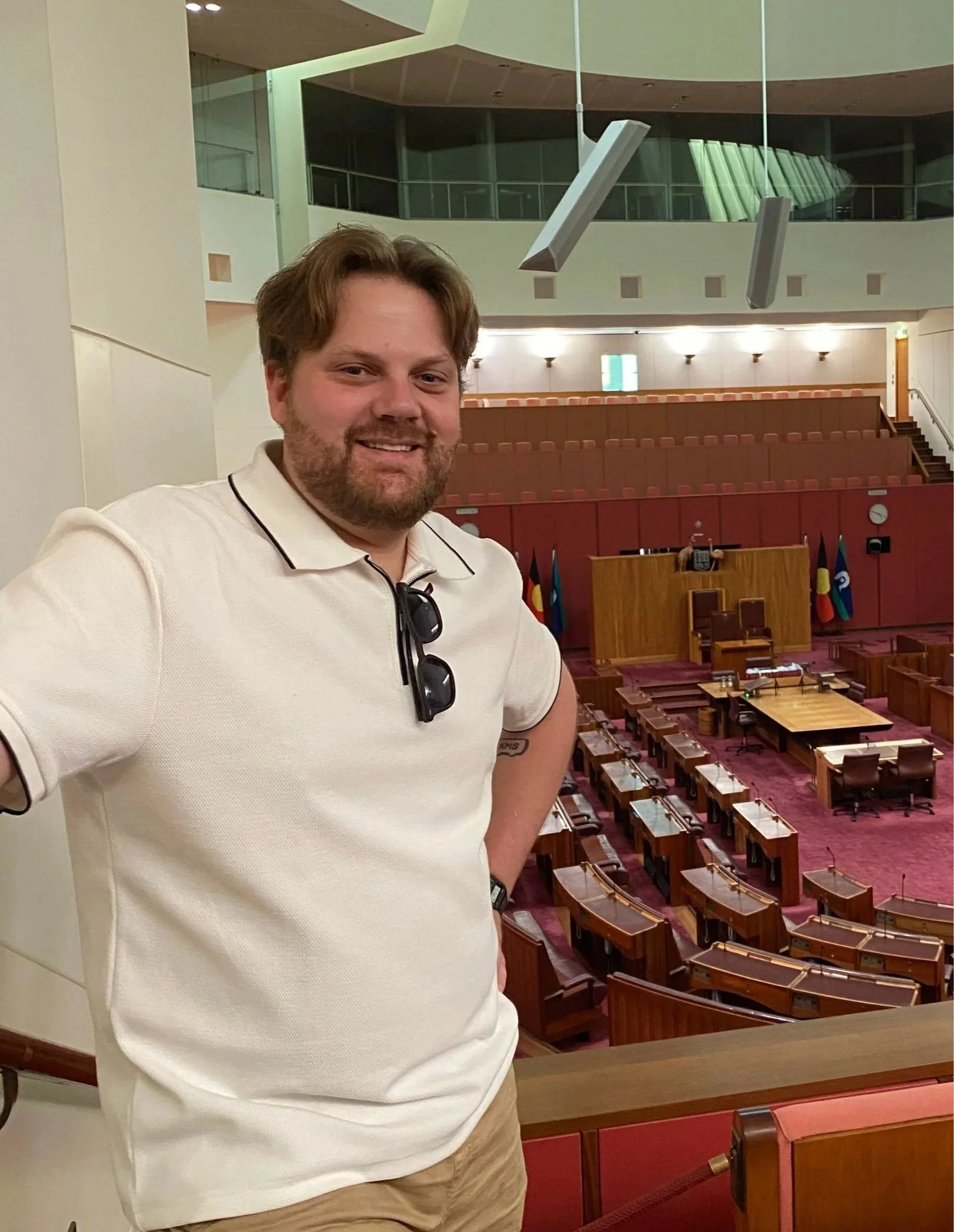 A man with a beard and brown hair smiling, standing inside a legislative chamber with a wooden podium and several flags.