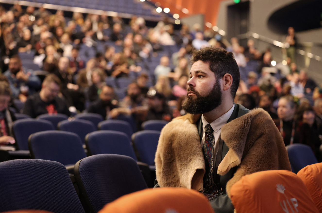 A man with a beard and dark hair, wearing a jacket with a fur collar, sitting alone in an auditorium filled with people, some of whom are clapping.