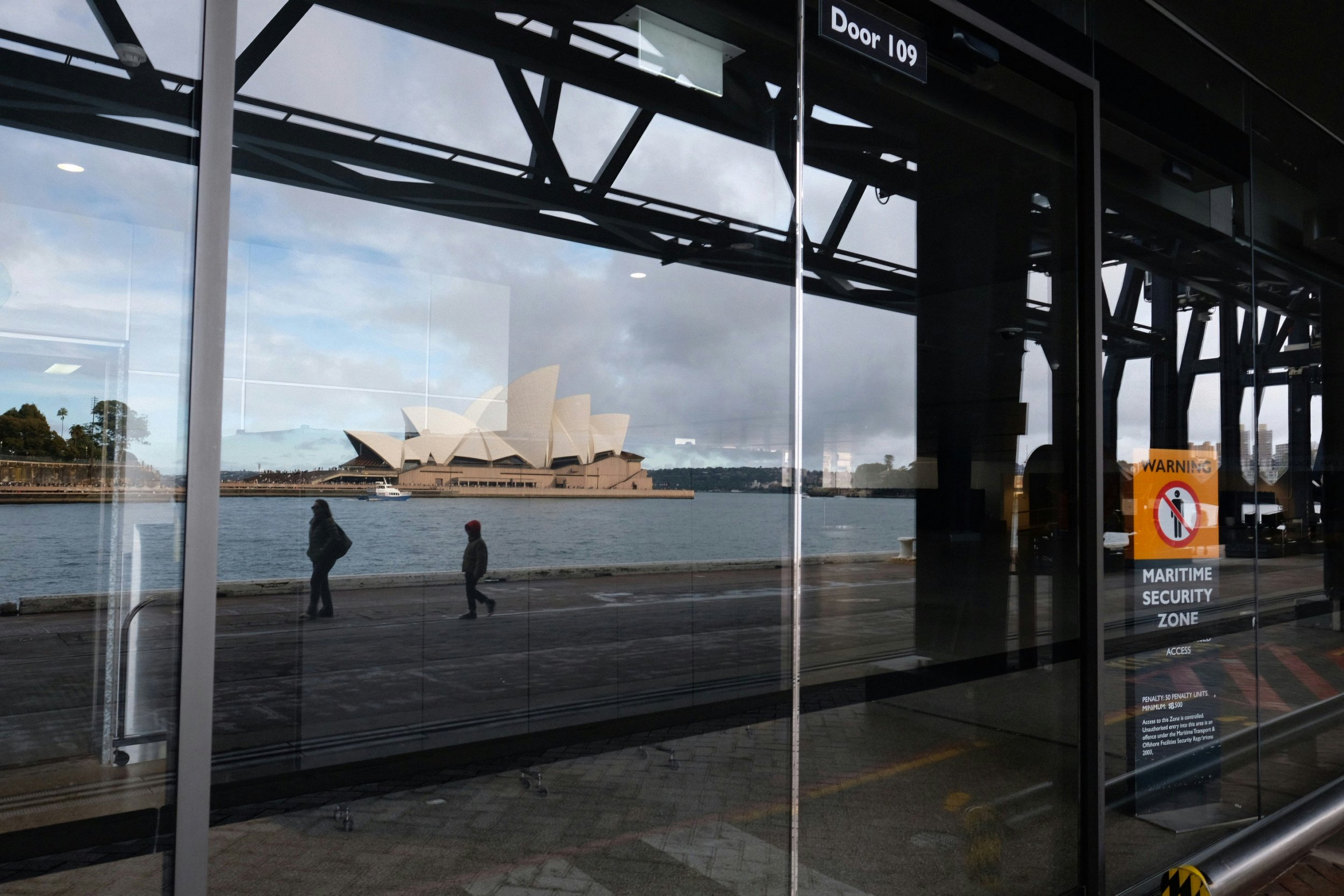 Reflection of the Sydney Opera House and two pedestrians walking along the waterfront seen through the glass doors of a maritime security zone.