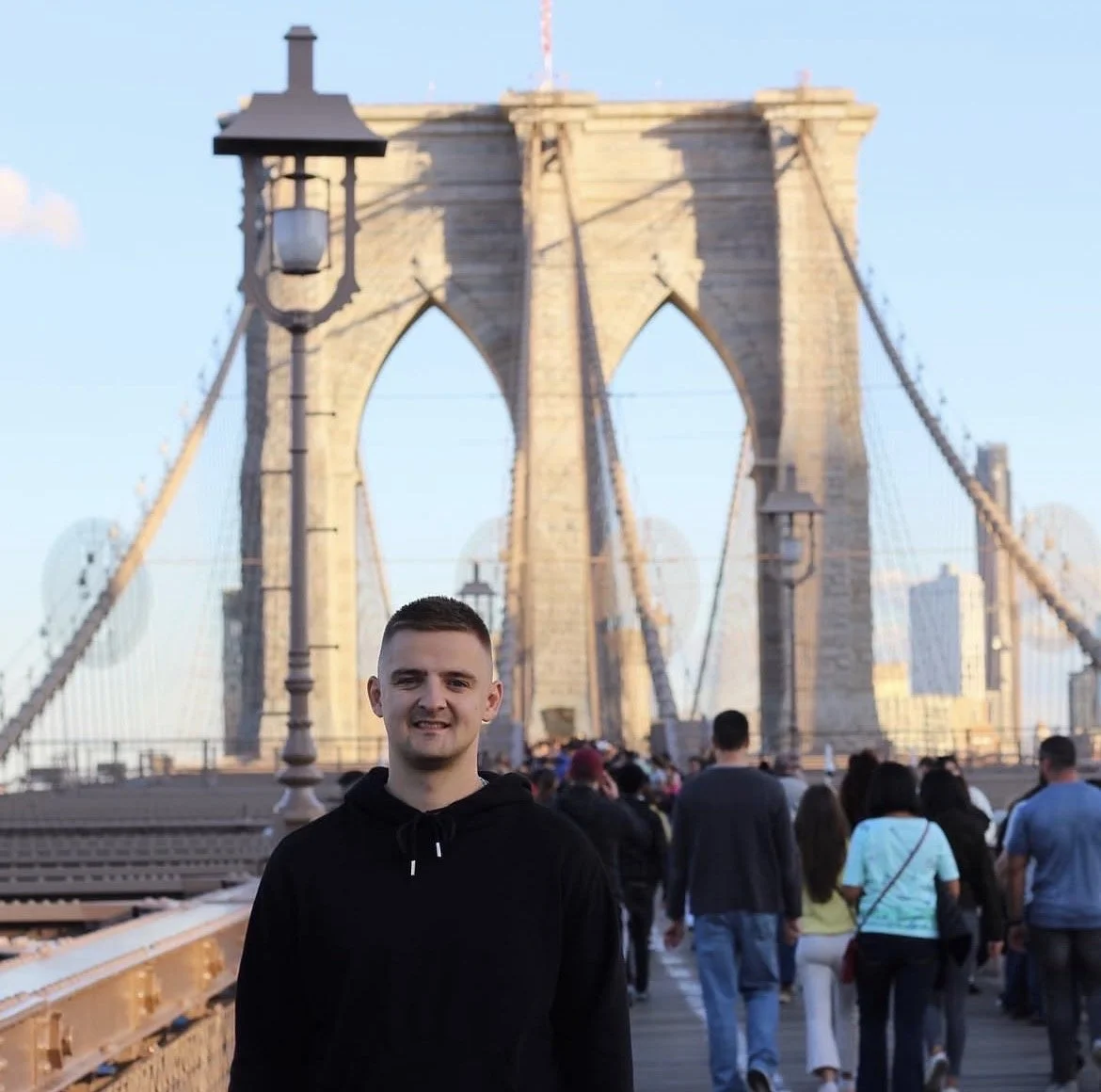 A young man in a black hoodie standing on the Brooklyn Bridge with the bridge's stone towers and suspension cables in the background, with a crowd of people walking across the bridge.