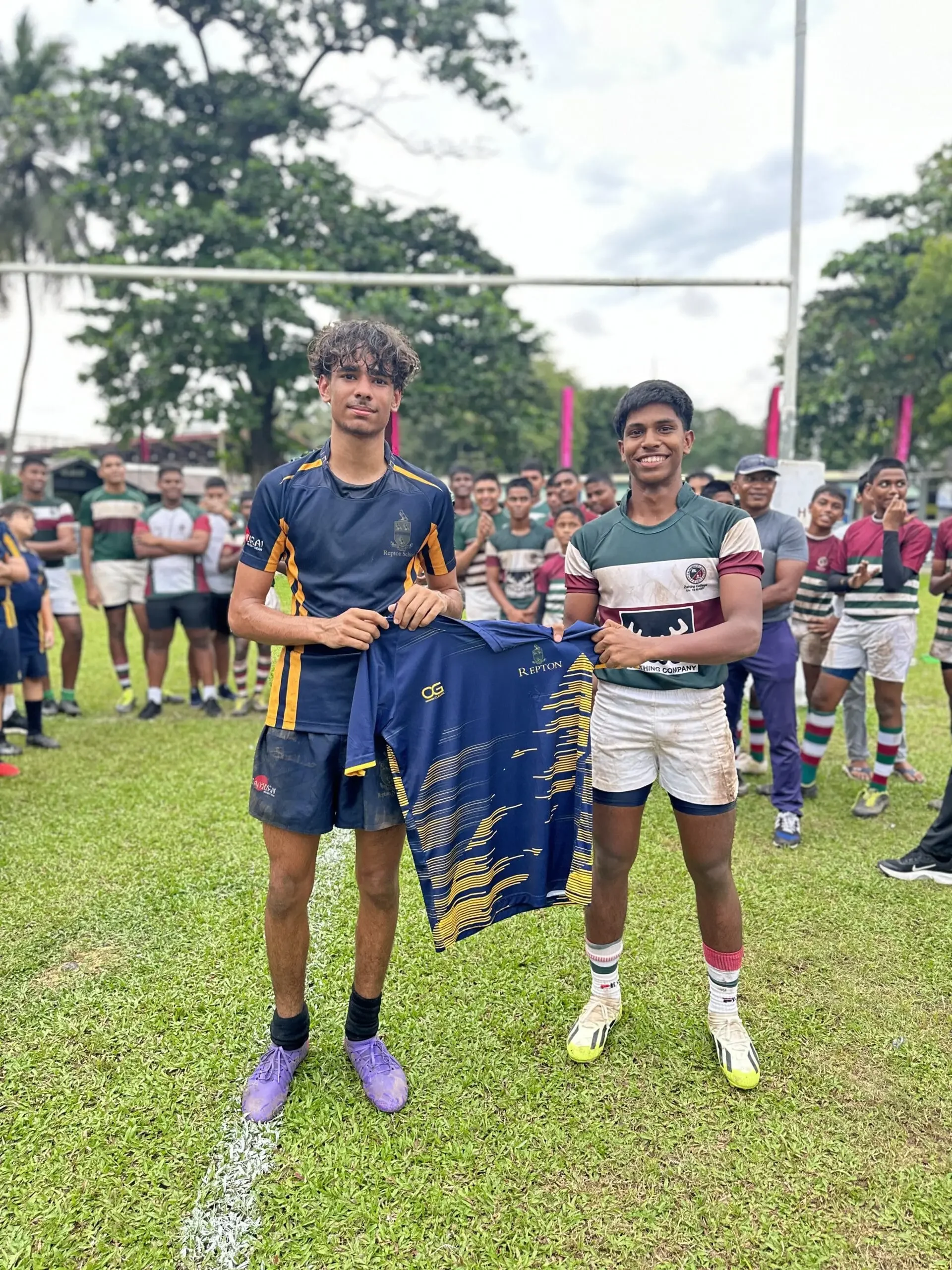 Two young men standing on a rugby field, holding a blue and yellow rugby jersey, with a large crowd and rugby posts in the background, surrounded by trees under a cloudy sky.