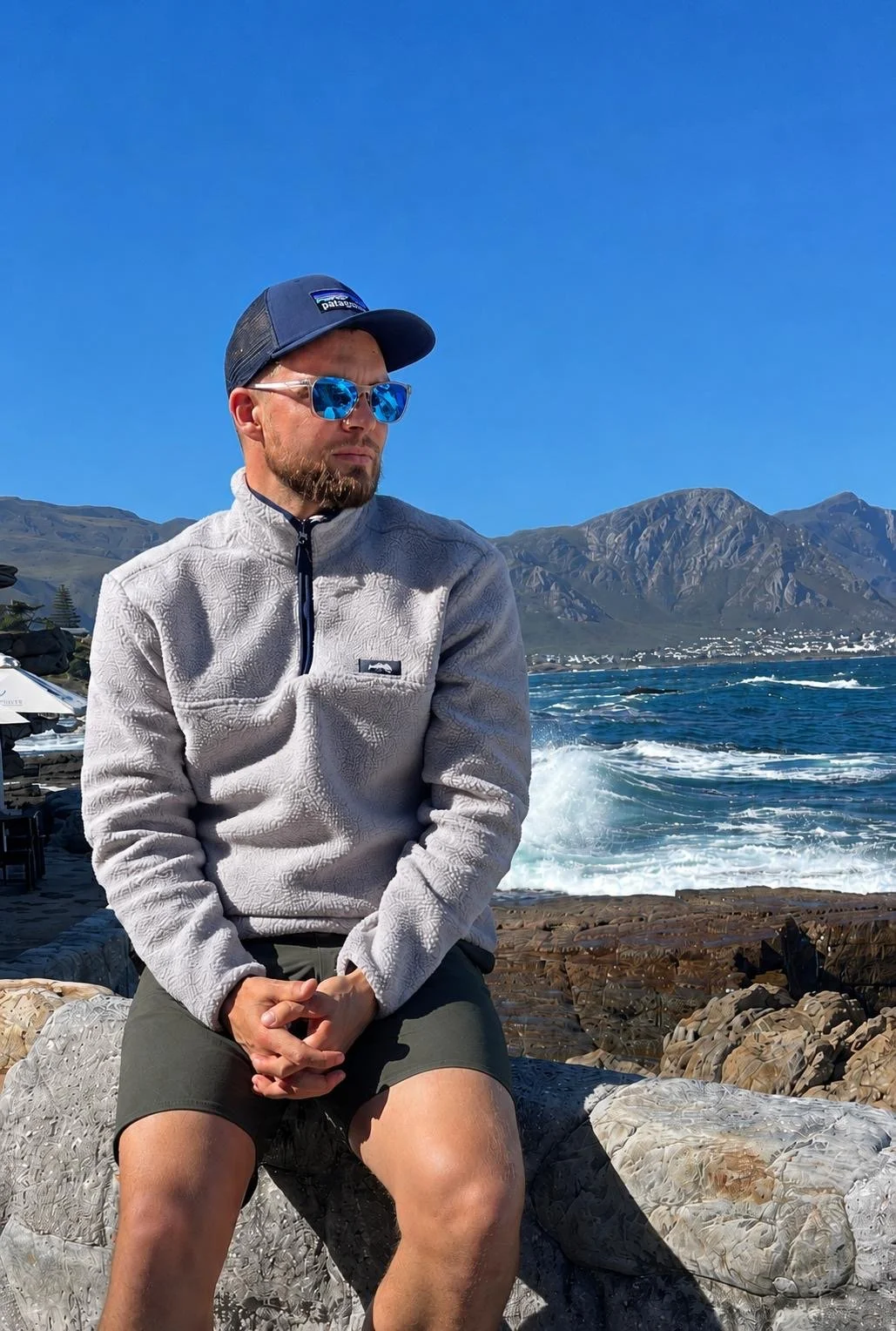 A man wearing sunglasses, a cap, and a light gray fleece jacket sits on rocks by the ocean, with mountains and a clear blue sky in the background.