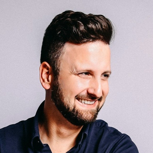 A man with dark hair and a beard smiling, wearing a dark shirt, against a light gray background.