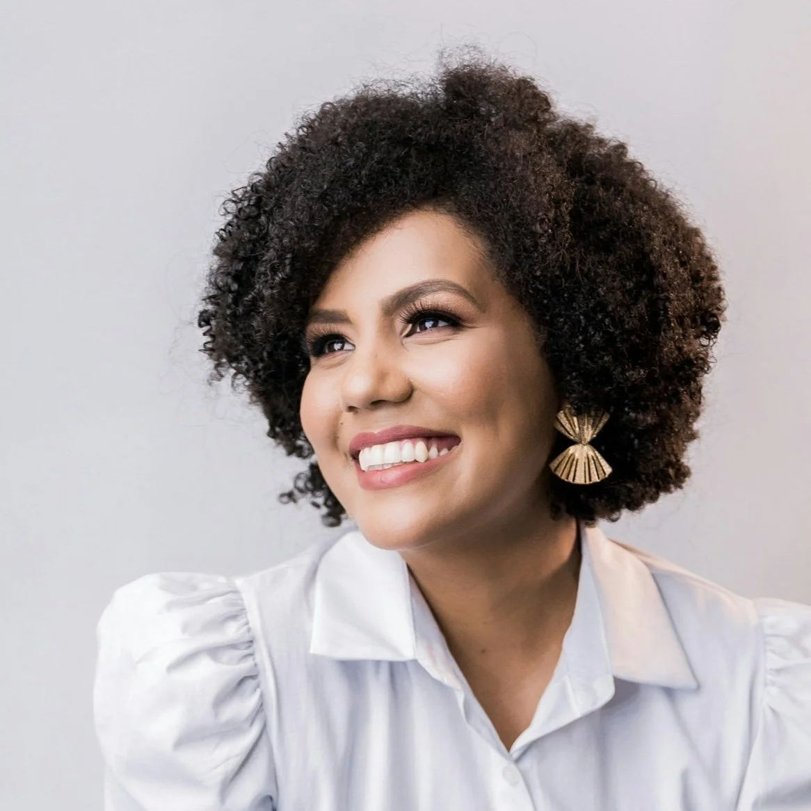 A woman with a big, natural curly hairstyle, wearing a white blouse and large gold earrings, smiling and looking to the side.