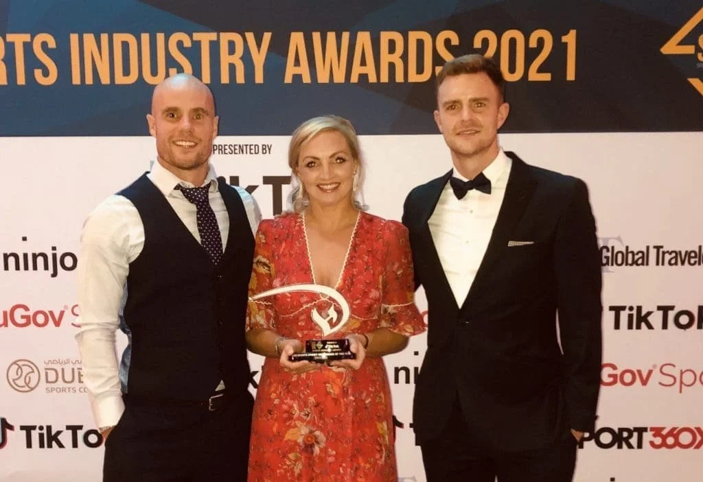 Three people dressed in formal attire at an awards ceremony, with a backdrop displaying 'Sports Industry Awards 2021' and various sponsor logos.