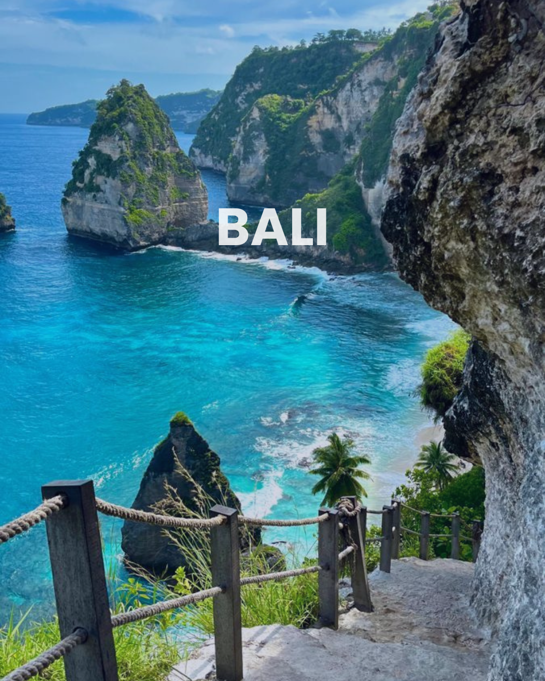 Coastal view of turquoise water and limestone cliffs in Bali, Indonesia, with a wooden railing along a path in the foreground.