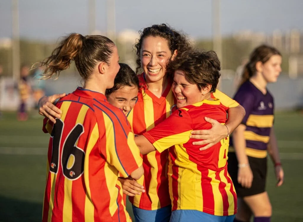 Four women's soccer players in red and yellow striped jerseys celebrate together on the field, with a woman wearing a purple and yellow striped jersey walking in the background.