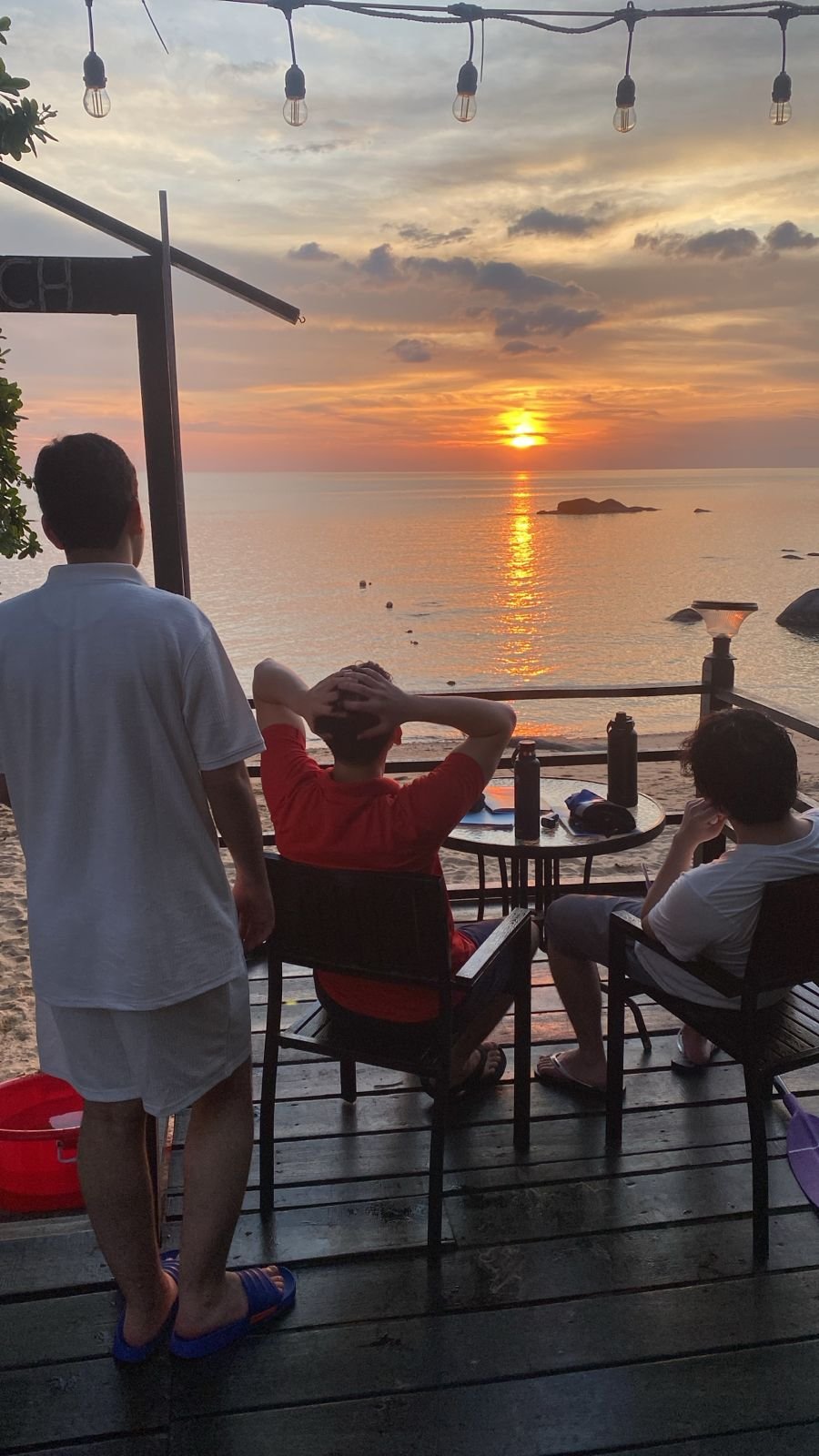 Three young men sitting and standing on a wooden deck overlooking the ocean during sunset, with string lights above and a table with water bottles in front of them.