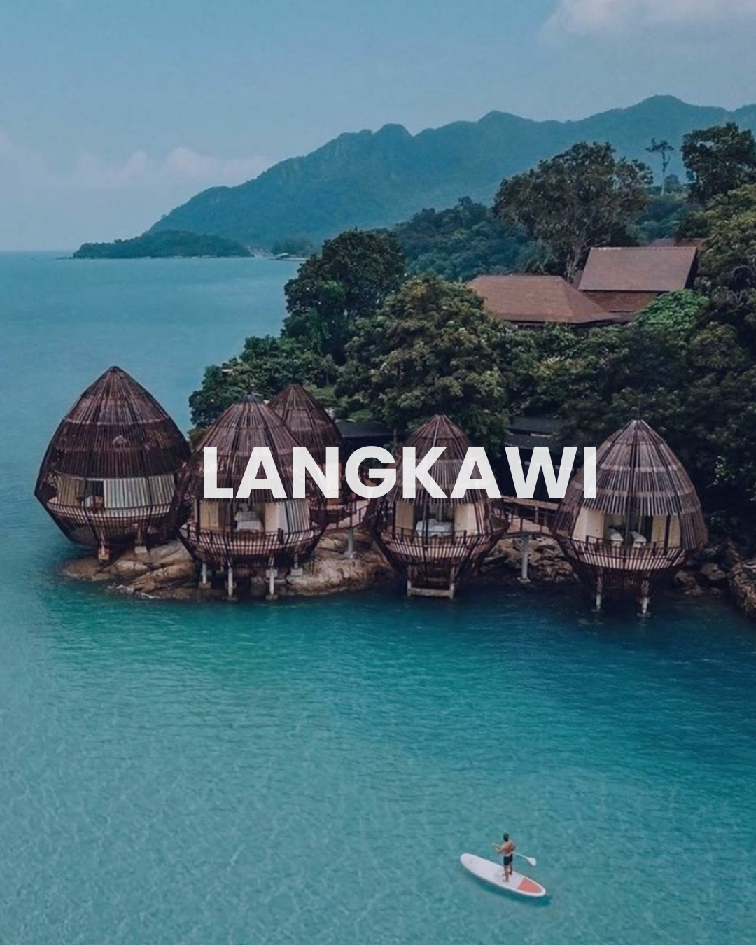 Overwater bungalows in Langkawi, Malaysia, with a man paddleboarding on turquoise water and lush green hills in the background.