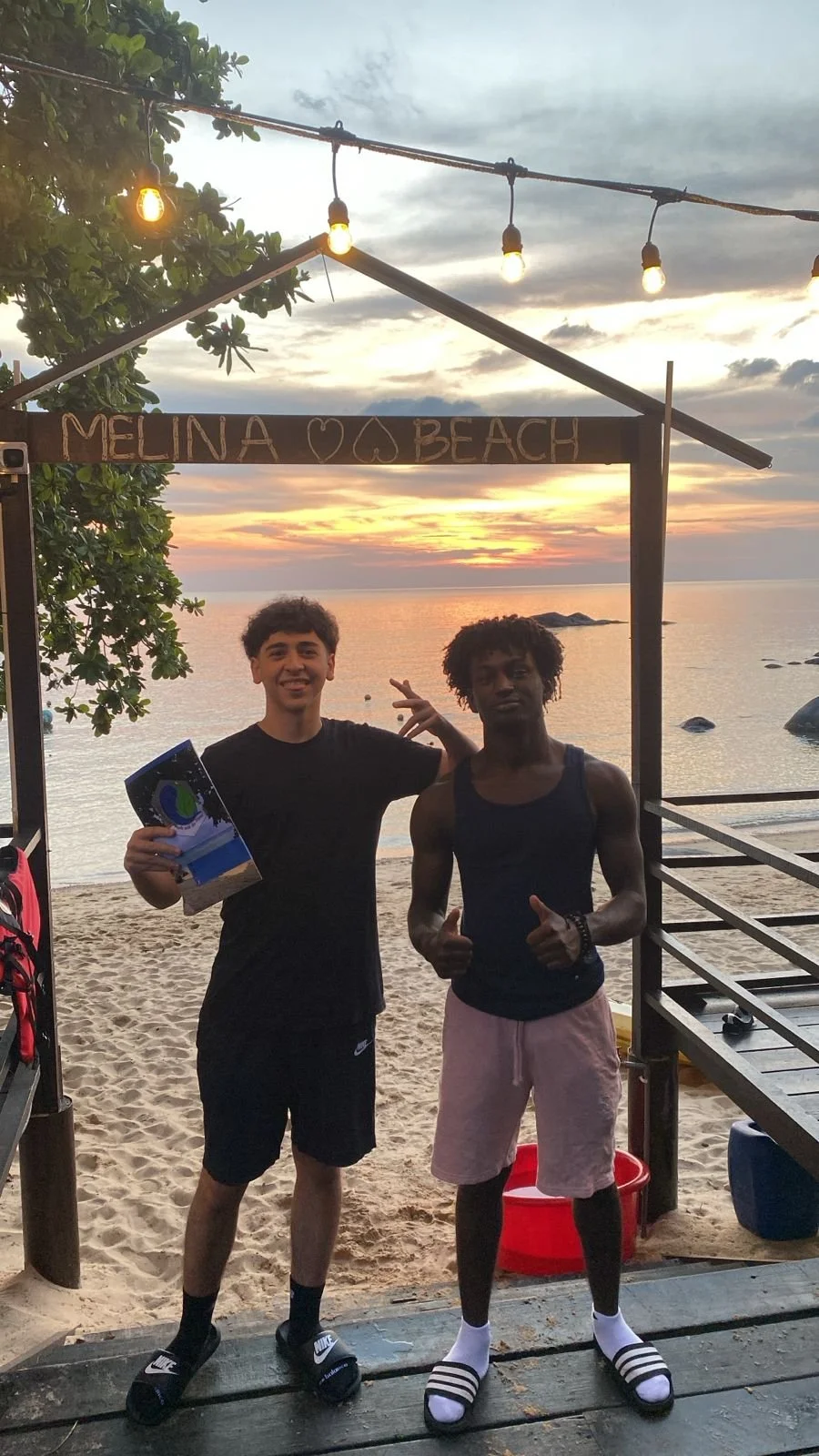 Two young men standing on the beach at sunset under a sign that reads 'Melina Beach,' with string lights overhead and the ocean in the background.