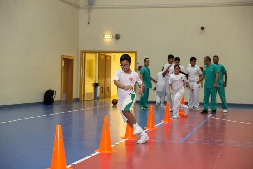 Children and healthcare workers participating in a physical activity session taking turns stepping over orange cones inside a gymnasium.