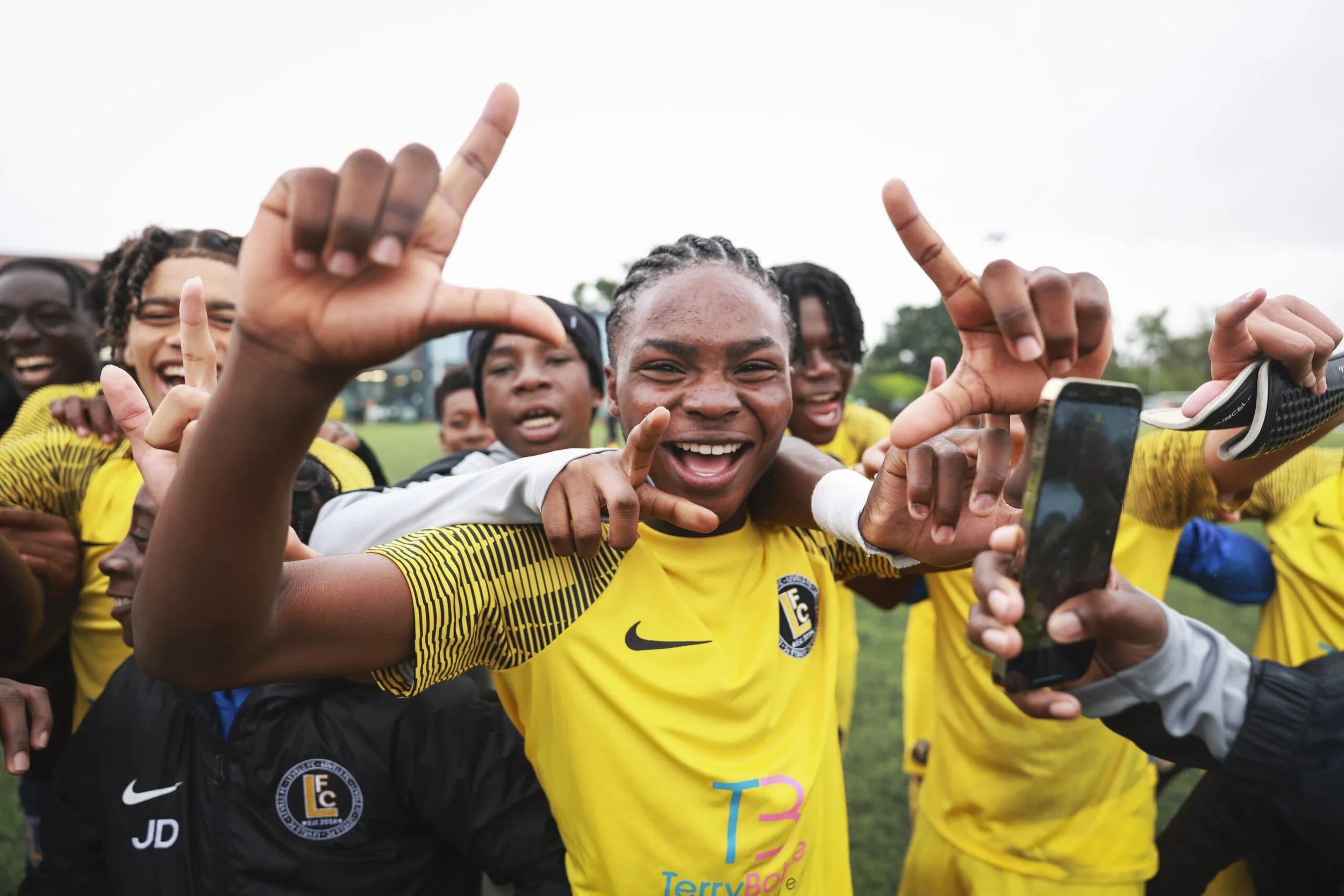 A group of smiling young soccer players wearing yellow jerseys huddle together for a selfie, with some making hand gestures and holding a phone, on a grassy field outdoors.