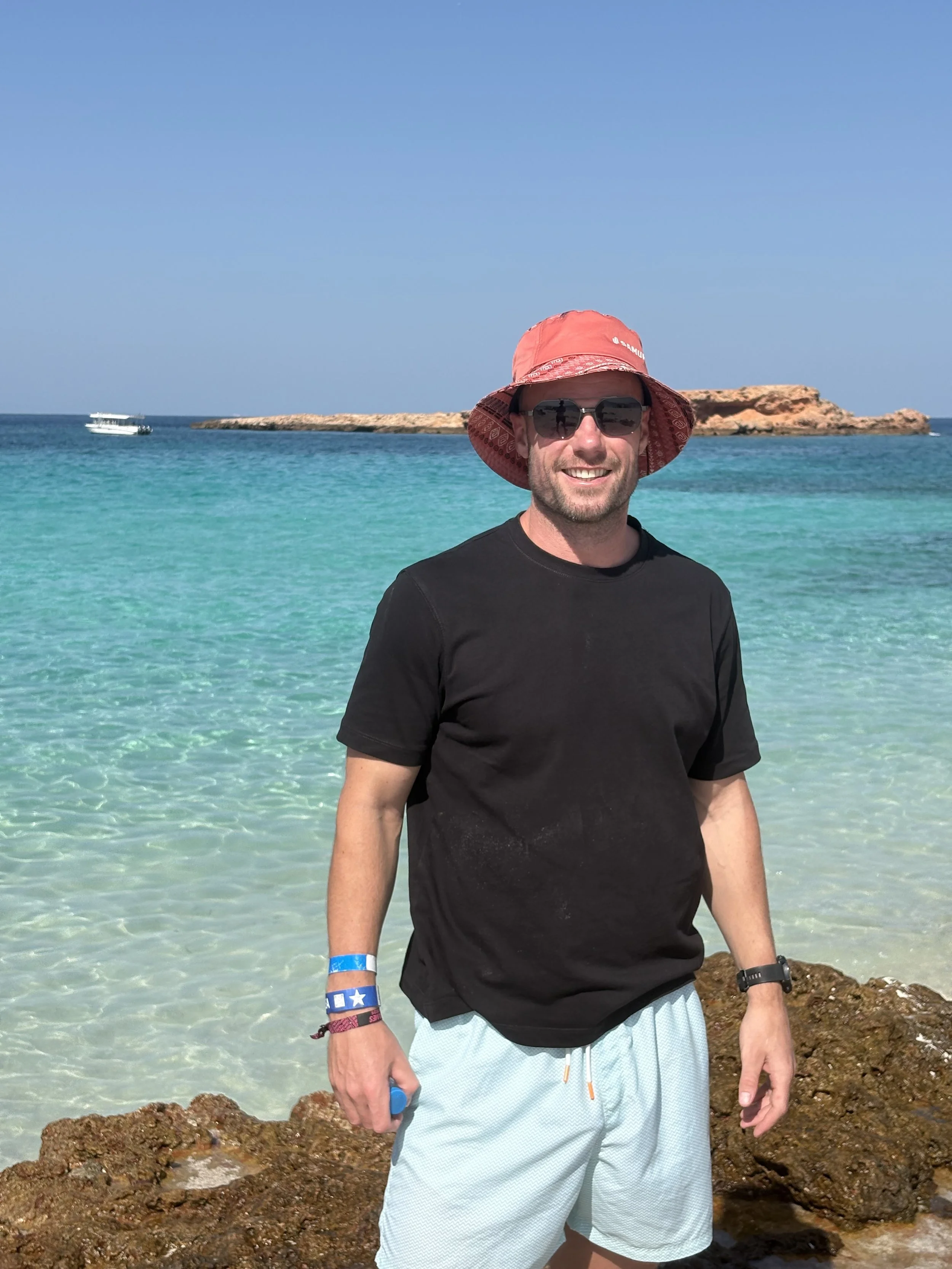 A man wearing a red wide-brimmed hat, dark sunglasses, a black t-shirt, and light-colored shorts standing on rocks by the ocean with clear blue water and small boats in the background.