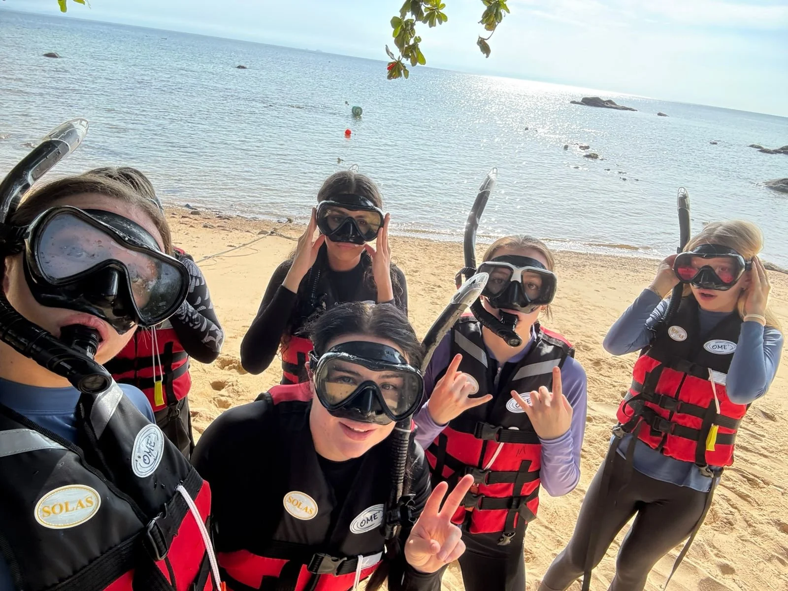 Six people wearing snorkeling gear and life vests on a sandy beach with the ocean and rocks in the background.