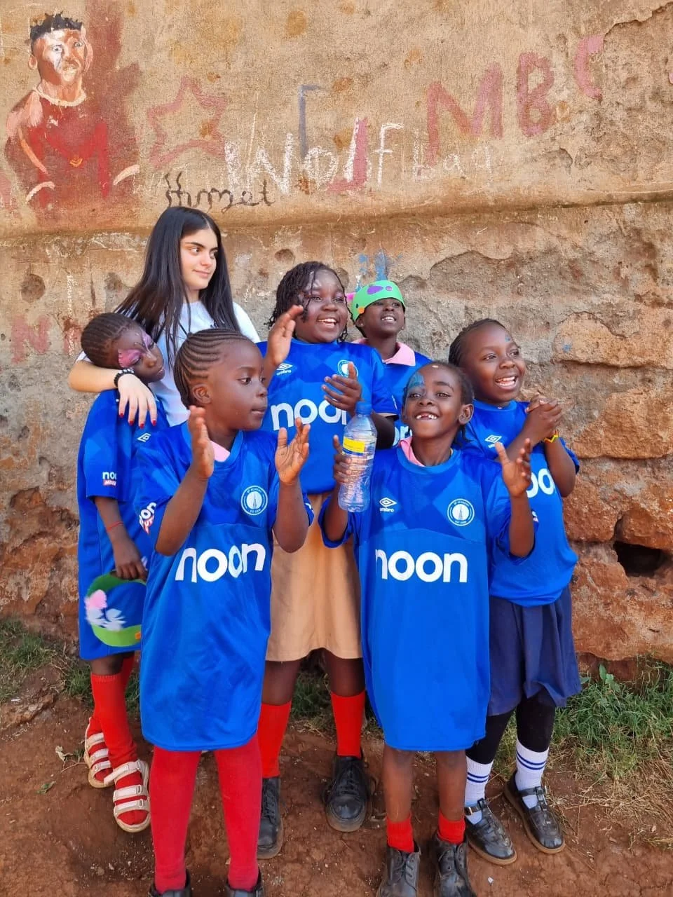 Group of children and a young woman standing outdoors against a rock wall, some children in blue sports jerseys with 'noon' logo, smiling and clapping, one holding a water bottle, an adult woman with long dark hair standing behind them.