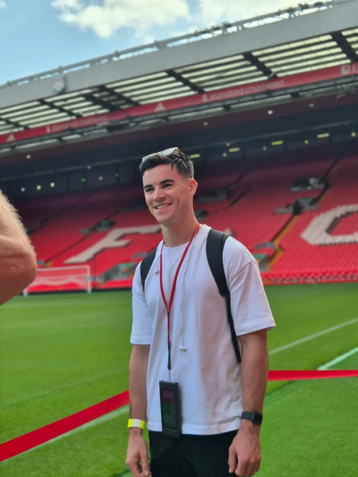 A young man with dark hair is standing on a soccer field in a stadium, smiling, wearing a white t-shirt, carrying a backpack, headphones around his neck, and a smartwatch on his left wrist.