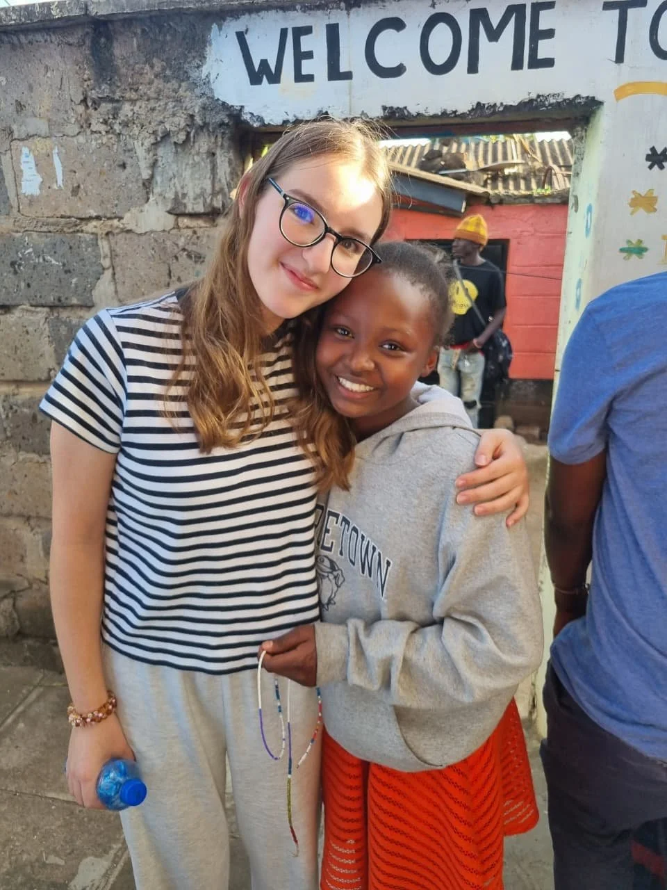 Two women, one with glasses and light skin wearing a striped shirt and sweatpants, and the other with darker skin wearing a gray hoodie and red skirt, hugging and smiling at the camera outside a building with a sign that says 'Welcome to'