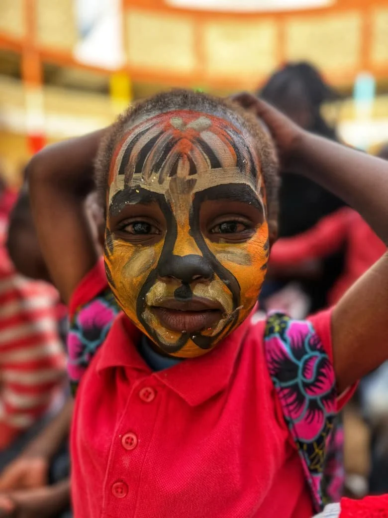 A young boy with face paint resembling a tiger, wearing a red polo shirt and standing at a colorful event or gathering.