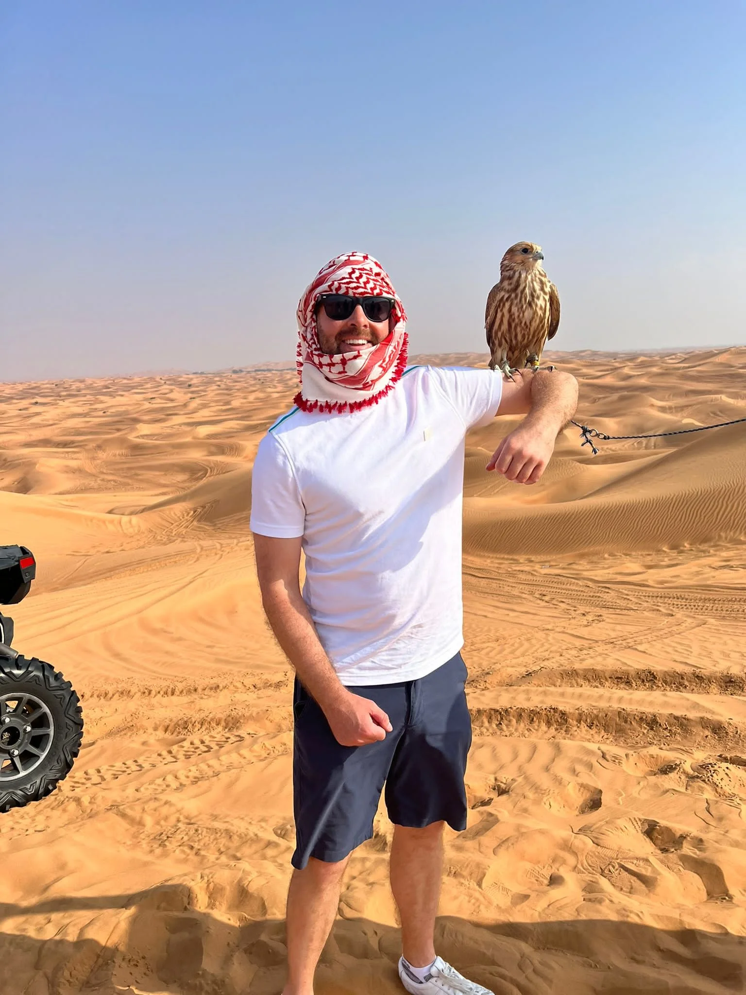 A man wearing sunglasses, a white t-shirt, and a red-and-white keffiyeh scarf, standing in a desert with sand dunes, holding a bird of prey on his arm.
