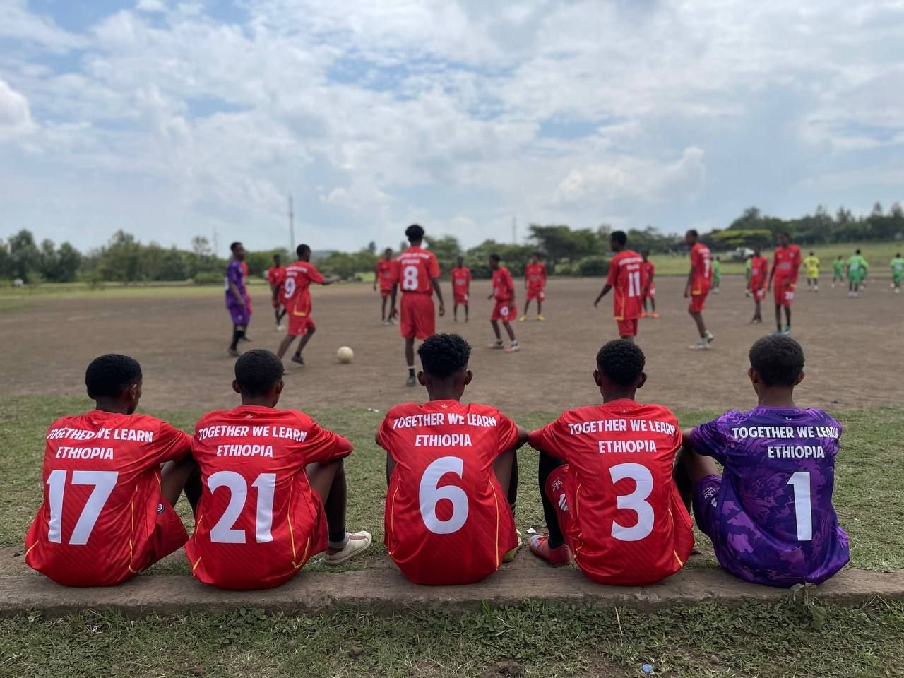 Ethiopian youth soccer team sitting on the sideline watching a game, with players on the field in red and green jerseys.