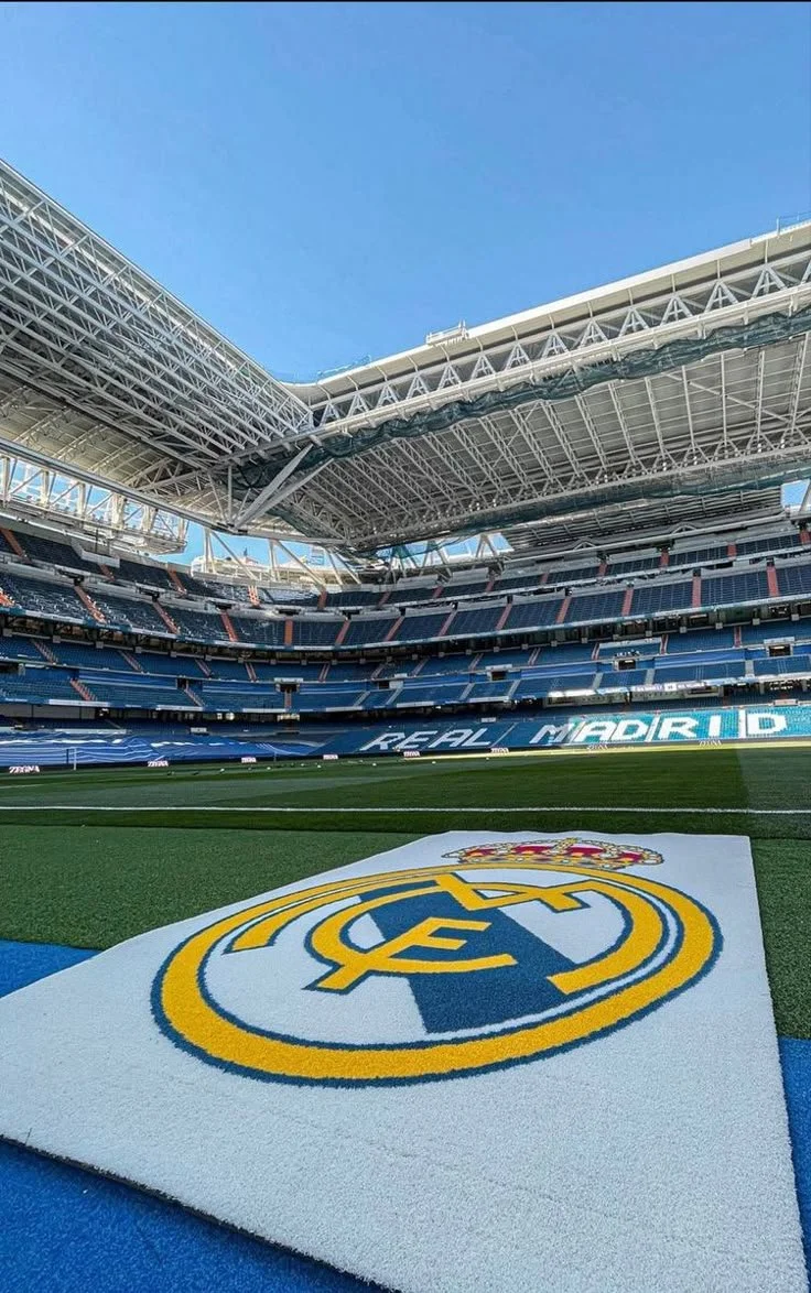 Inside a soccer stadium with a large Real Madrid emblem on the field and empty blue seats in the stands.