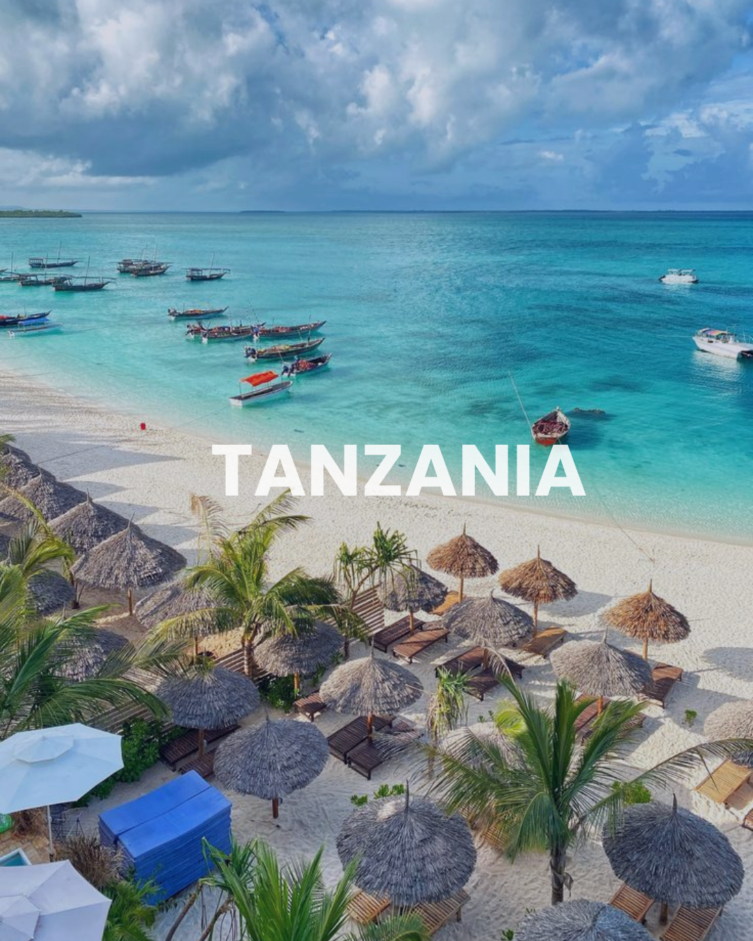 Tanzania beach with boats in turquoise water, sandy shore with thatched umbrellas, palm trees, and cloudy sky.