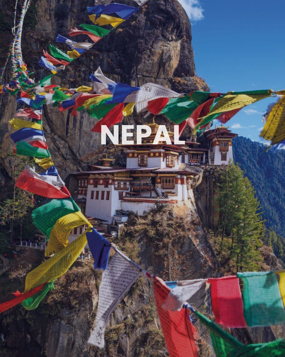 Colorful prayer flags flutter in front of a monastery built into a rocky cliff in Nepal, with mountains and blue sky in the background.