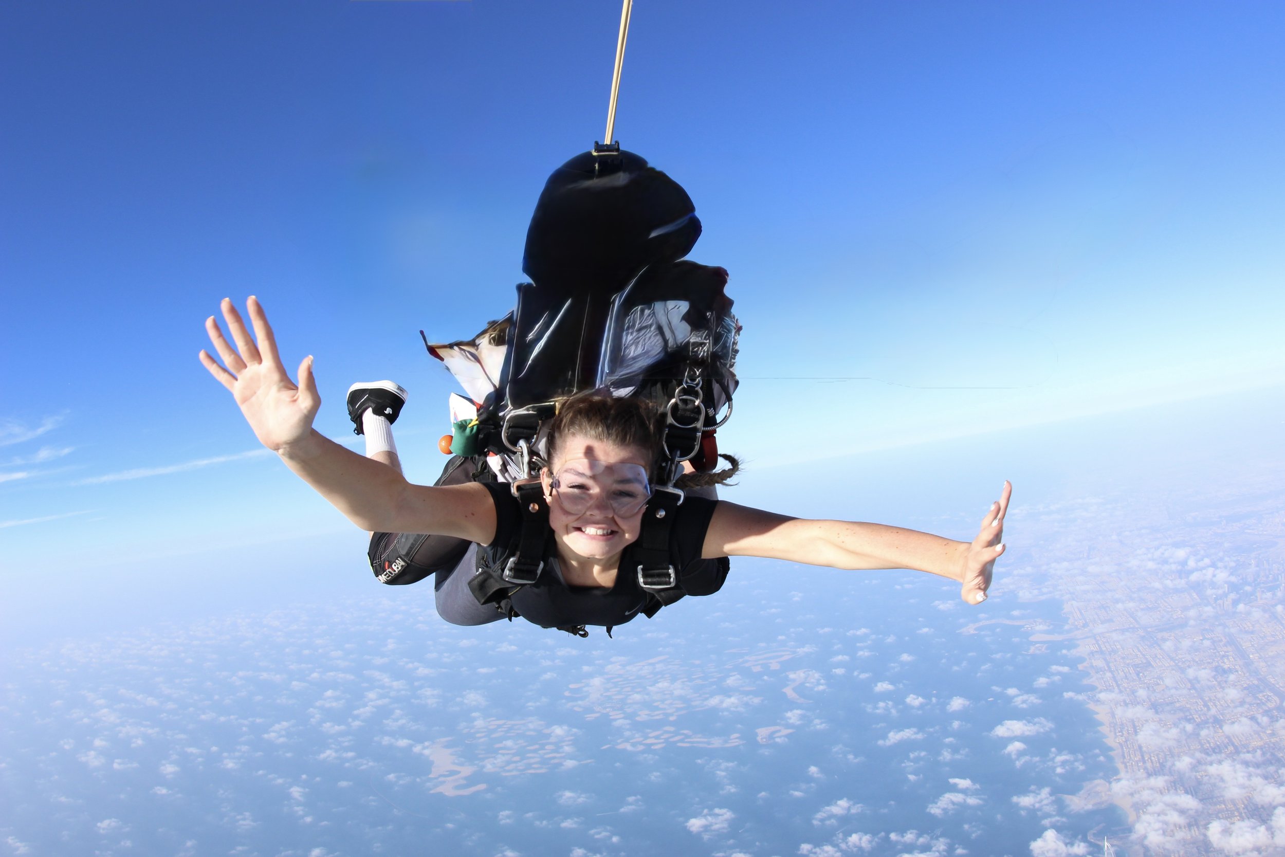 A woman skydiving outdoors with a wide smile, wearing protective goggles, with a sky and clouds in the background.