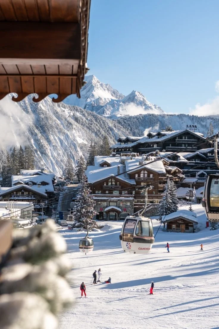 Snow-covered village with ski gondolas in the foreground, ski slopes with skiers, chalet-style buildings, and snow-capped mountains in the background on a clear day.