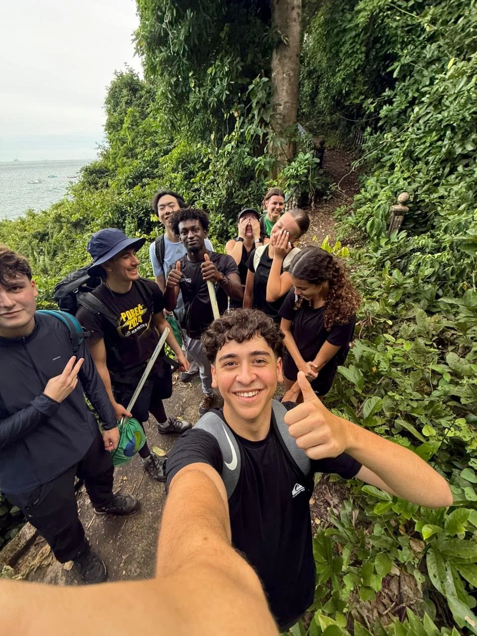 Group of people on a hiking trail in a lush, green forest near a body of water, taking a selfie.