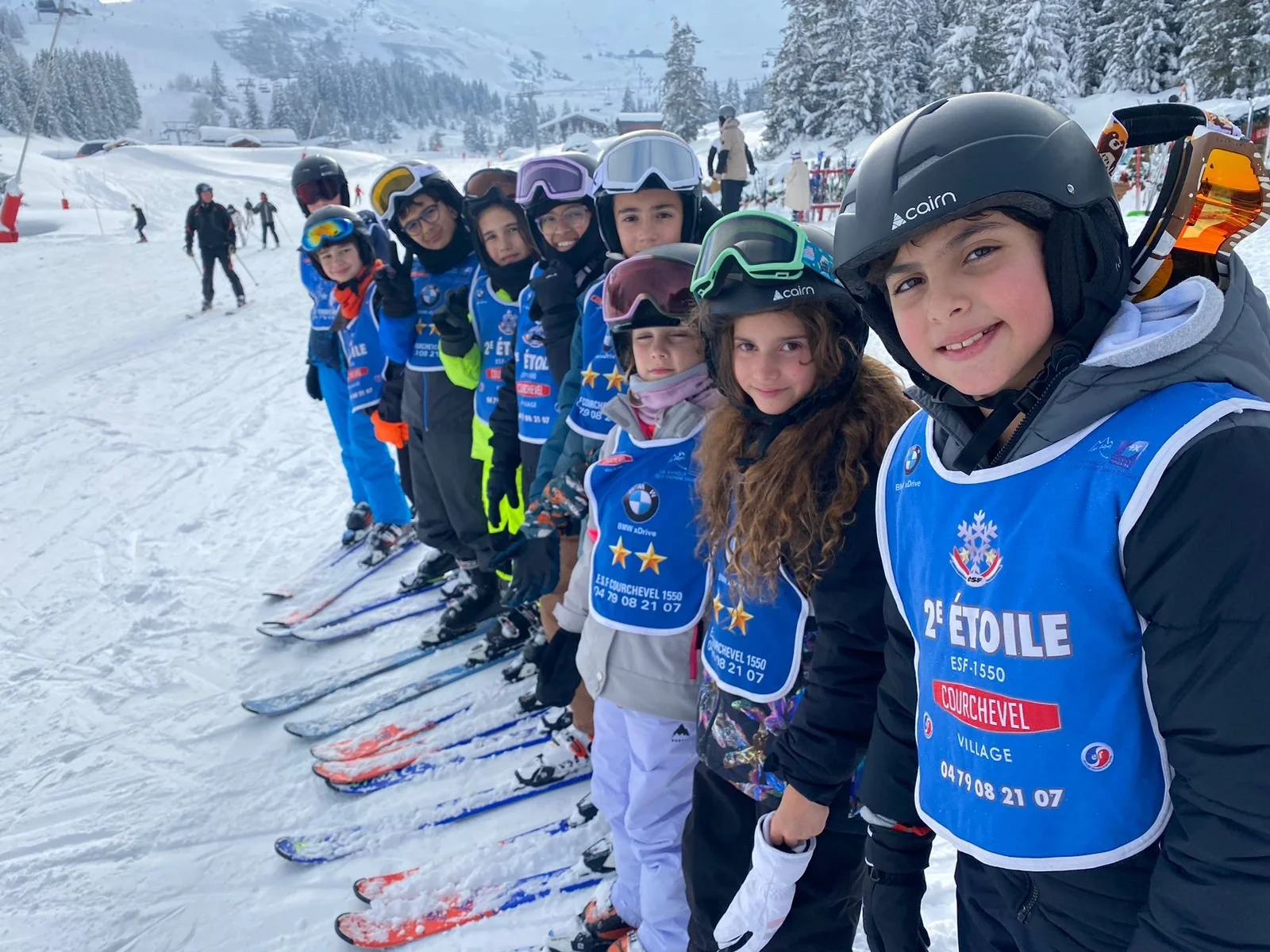 A line of children wearing ski gear and helmets, standing in snow-covered landscape with ski slopes and trees in the background, preparing for skiing.