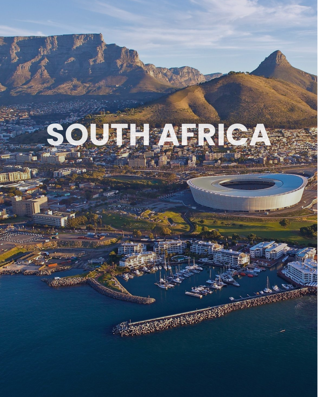 Aerial view of Cape Town, South Africa, with the city, harbor, mountains, and a stadium under a partly cloudy sky.