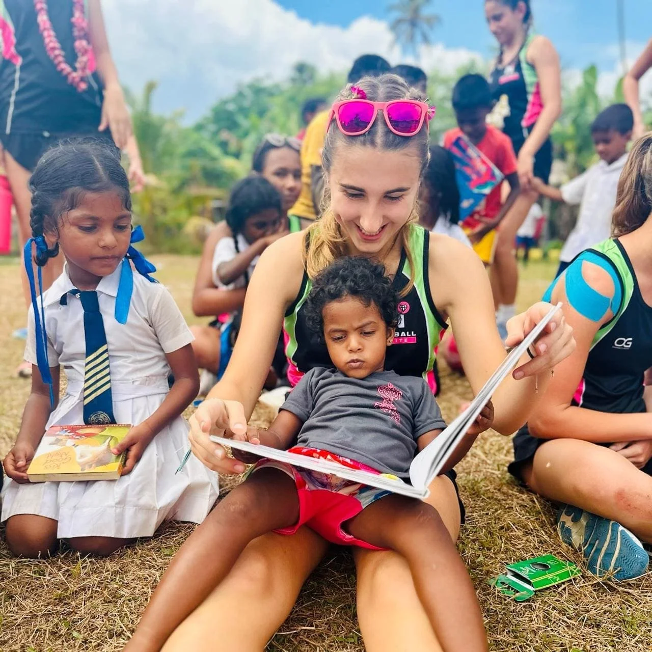 A group of children and a young woman reading a book outdoors on the grass, surrounded by greenery and a cloudy sky, during daytime.