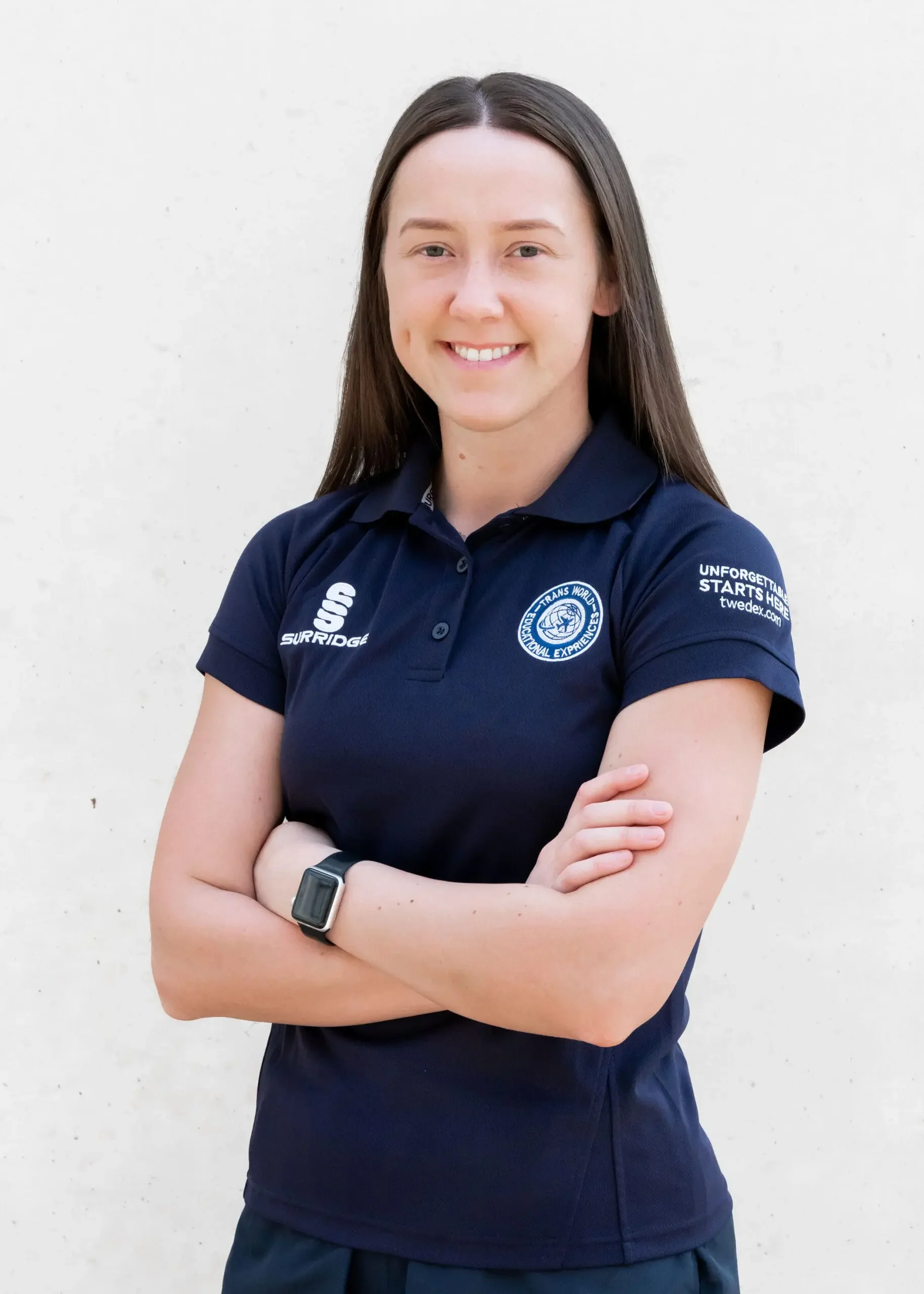 A woman with long brown hair smiling, wearing a navy blue polo shirt with logos, crossed arms, standing against a plain white background.