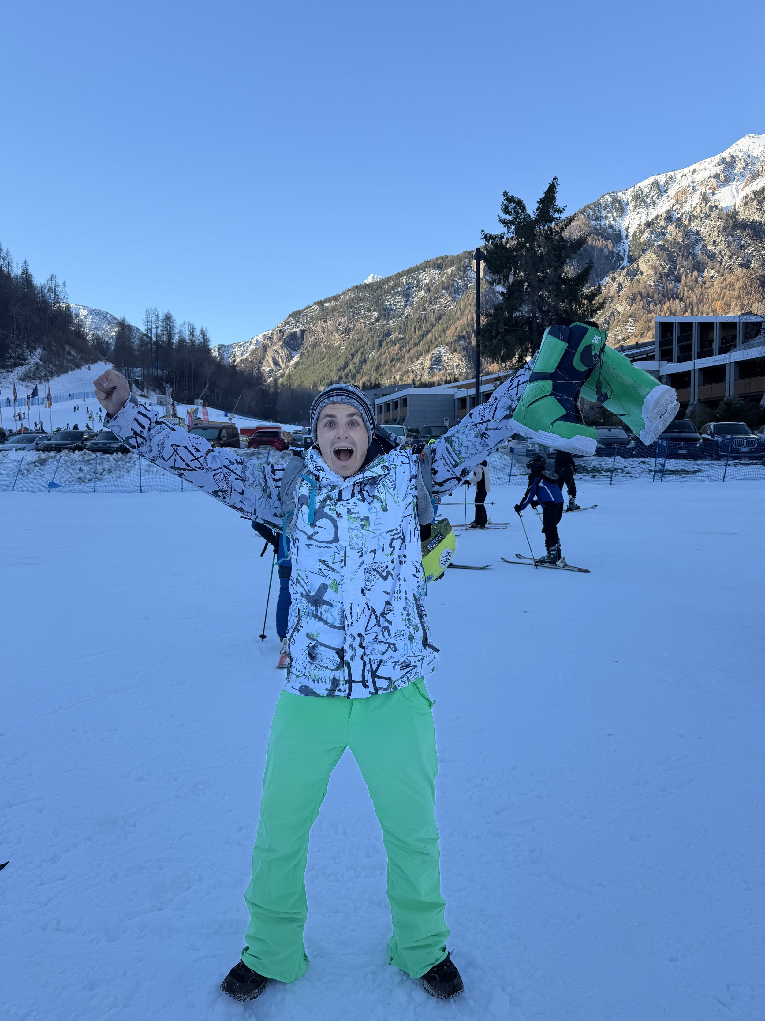 Excited man in colorful winter clothes celebrating at a snowy mountain ski resort with mountains, snow, and other skiers in the background.