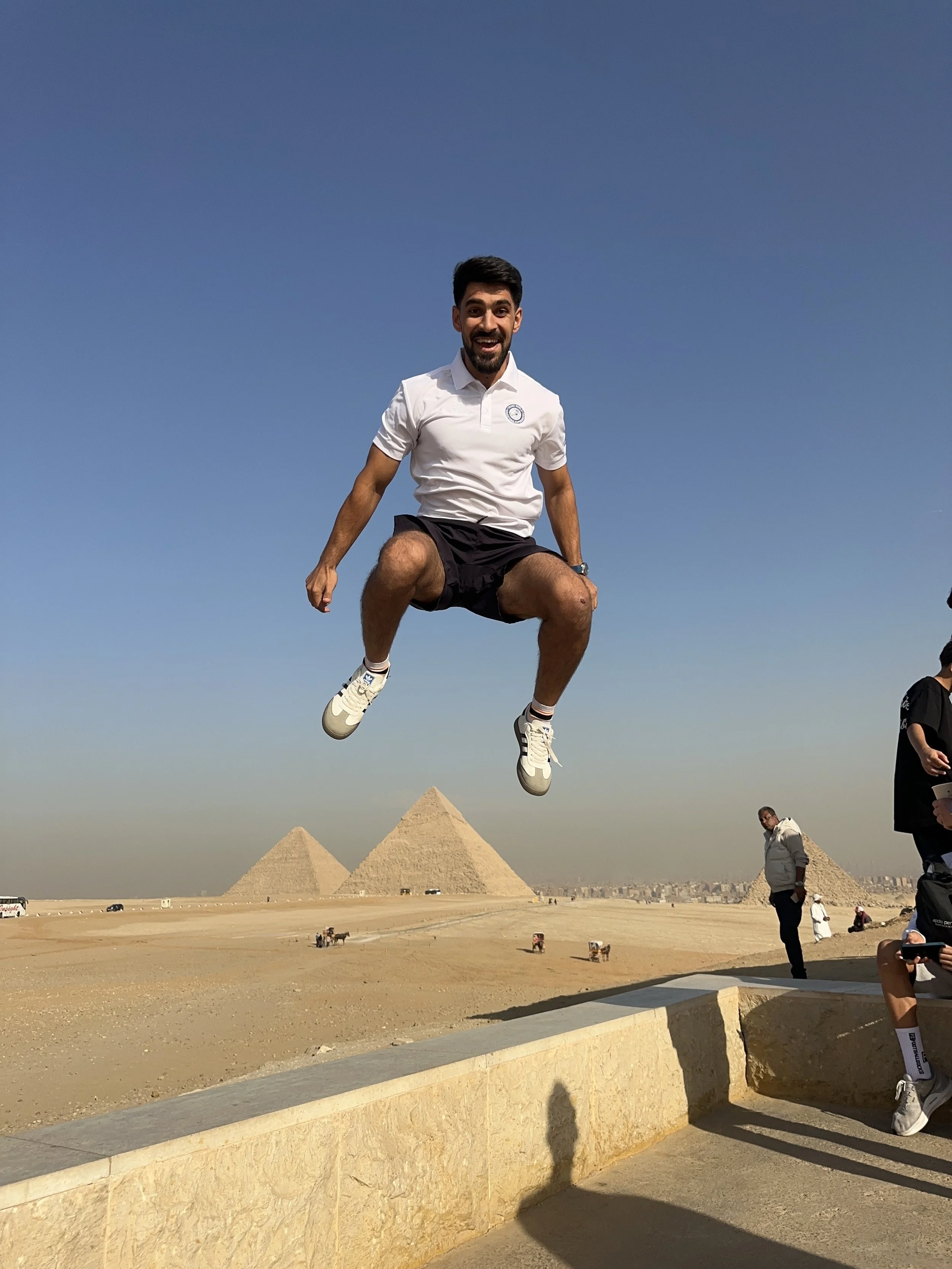 A man in a white shirt and black shorts jumps in the air with a big smile, with the pyramid of Khafre in the background on a clear day.