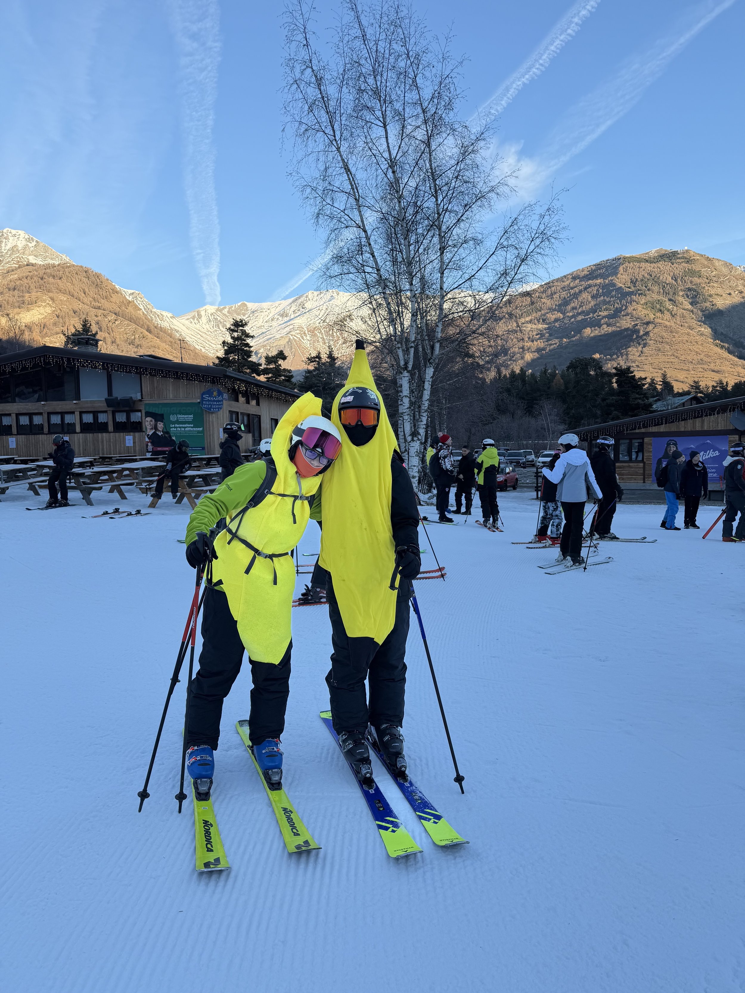 Two people dressed in bright yellow ski suits and helmets stand on a snow-covered ski slope, smiling. In the background, there are other skiers, a wooden building, trees, and mountains under a clear blue sky.