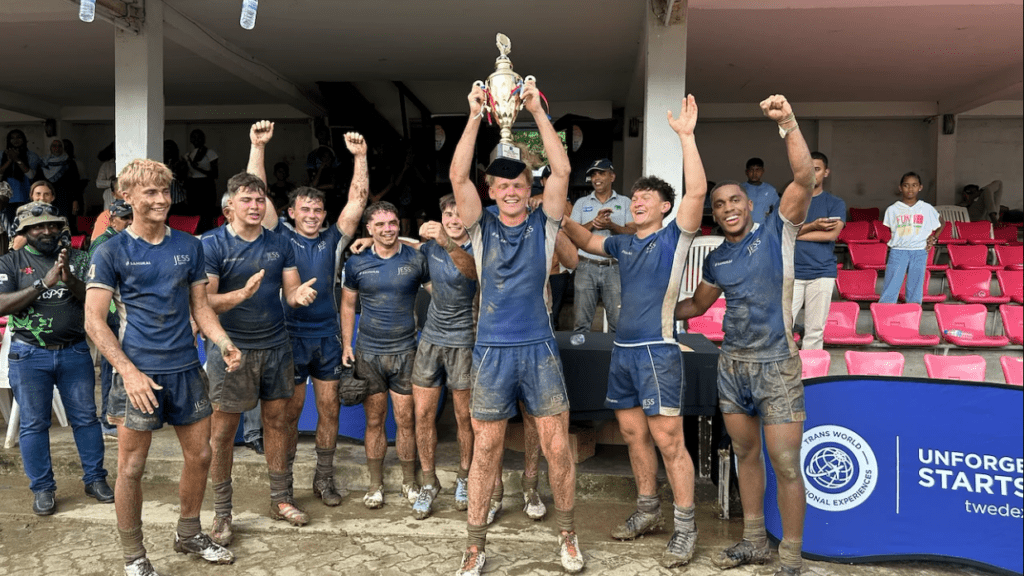 A group of young rugby players celebrating after winning a match, holding a trophy, and smiling. They are wearing muddy jerseys and shorts, standing on a muddy field.