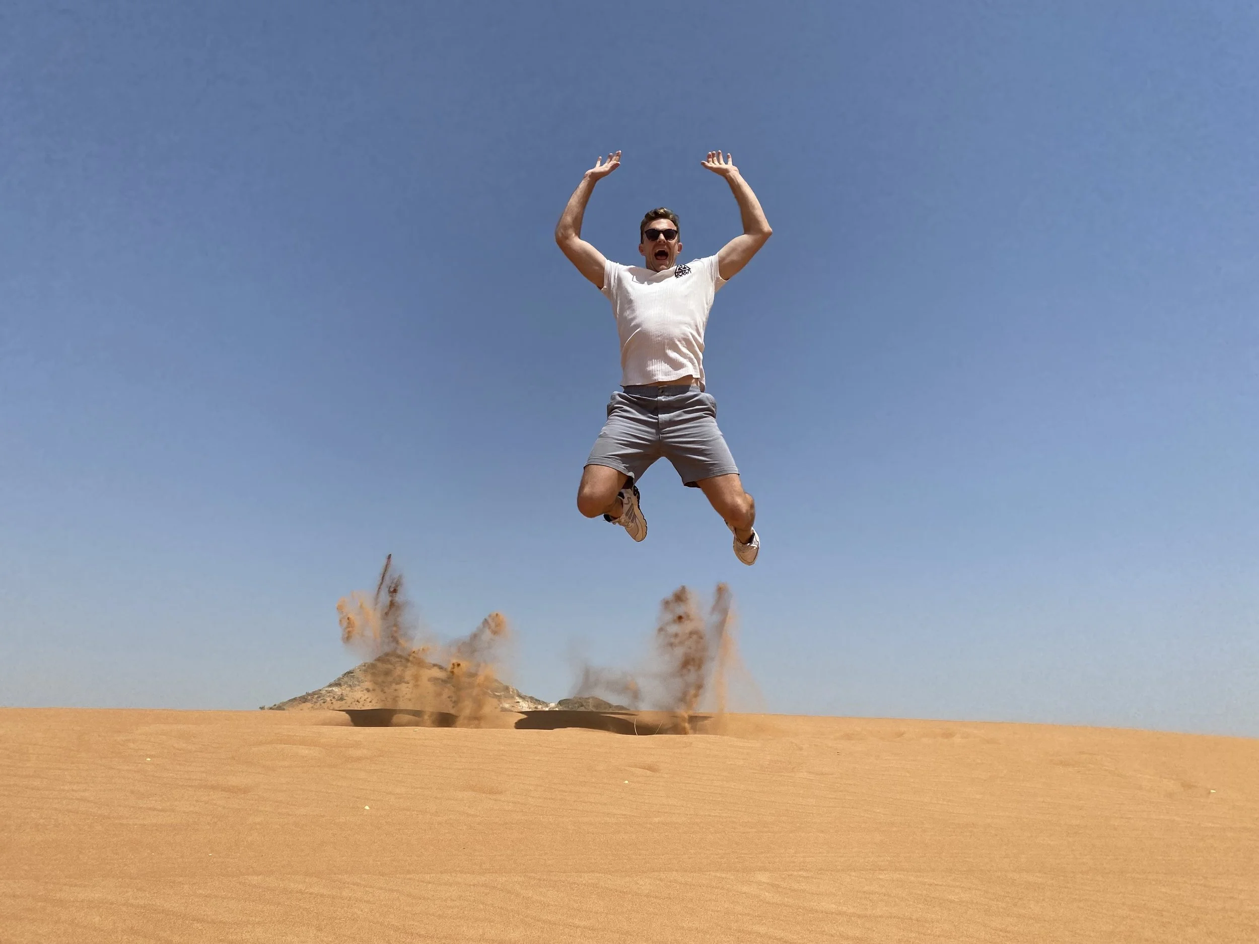 A man wearing sunglasses, a white t-shirt, and shorts jumping in the air with arms raised and mouth open in front of a desert landscape with mountain and blue sky.