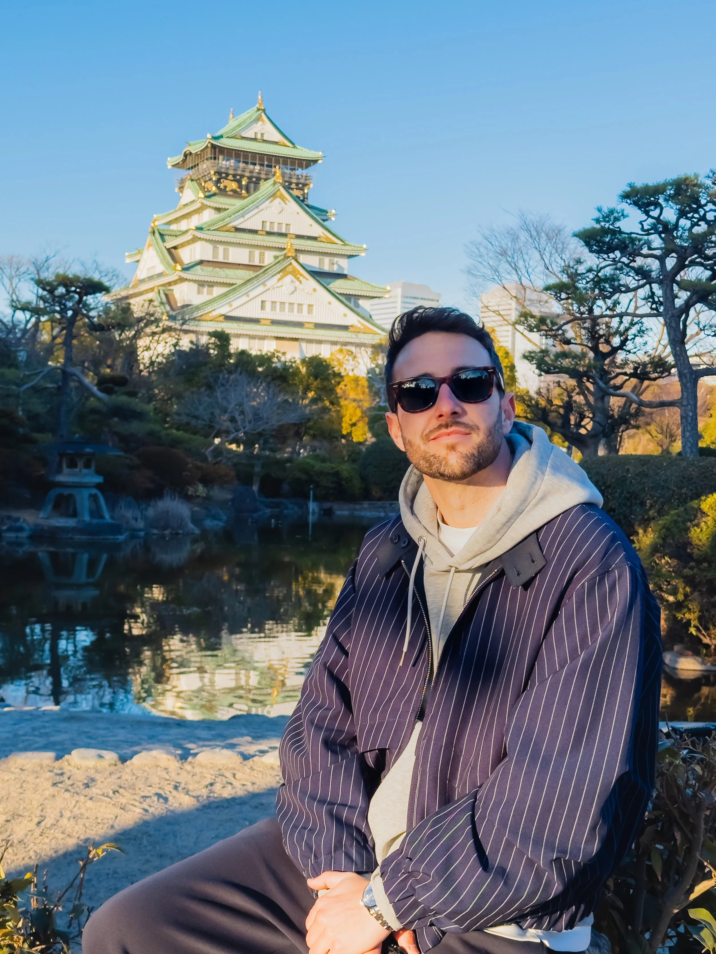 A man wearing sunglasses, a striped blazer, and a gray hoodie sitting by a pond with a traditional Japanese castle in the background during daytime.