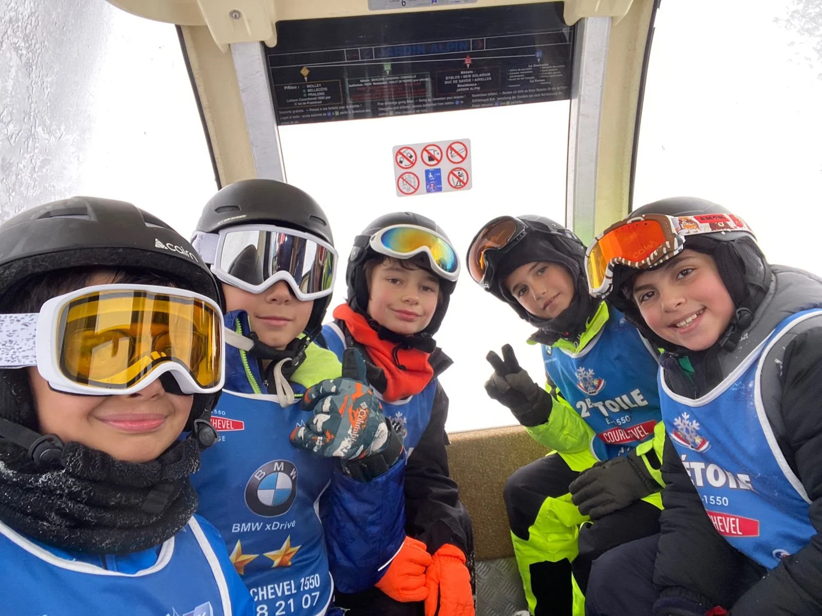 Five young skiers wearing helmets and goggles taking a selfie inside a ski lift. They are dressed in blue ski uniforms with sponsor logos, smiling and showing peace signs.