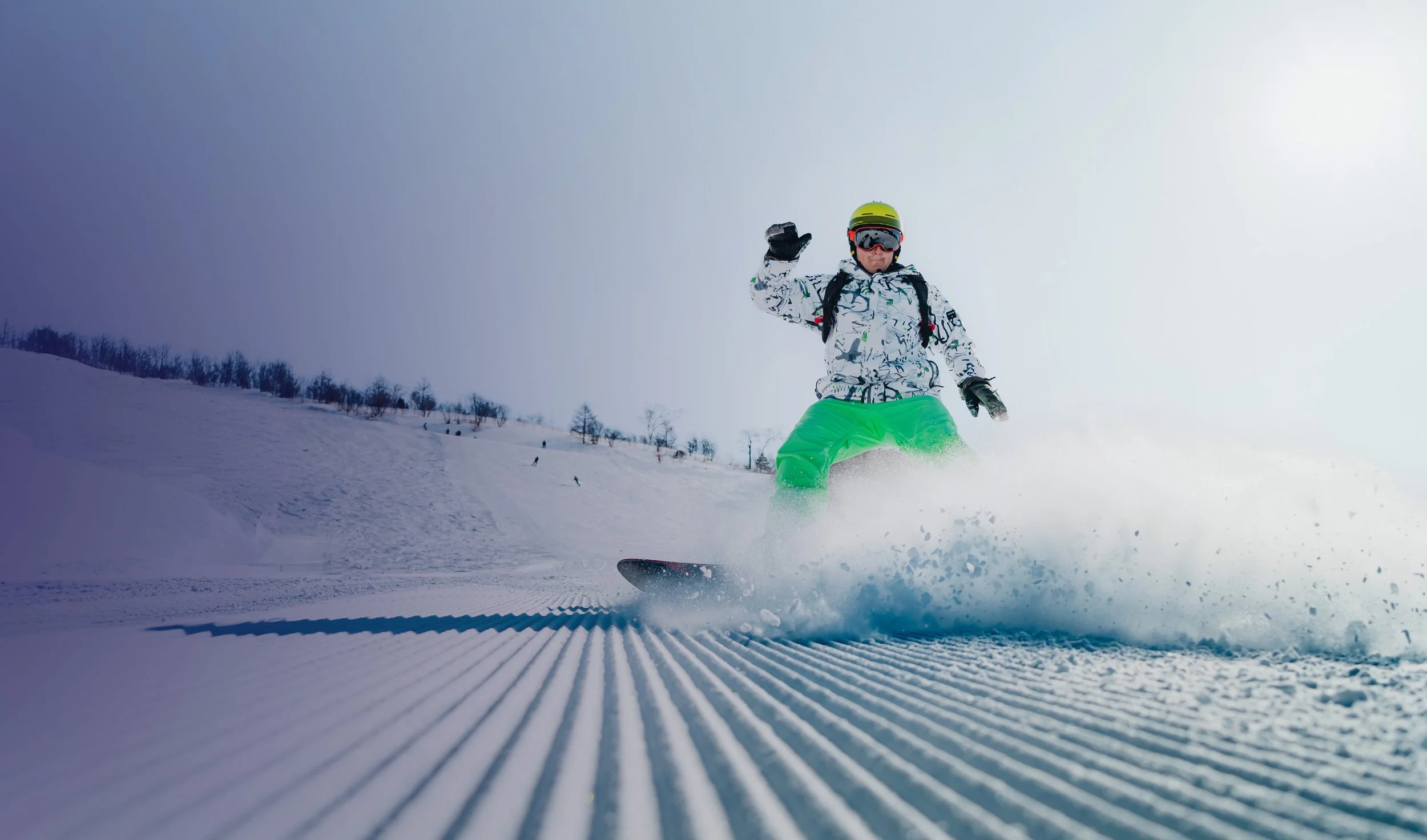 A snowboarder wearing a white patterned jacket, bright green pants, a yellow helmet, and goggles, is gliding down a snow-covered slope with trees in the background.