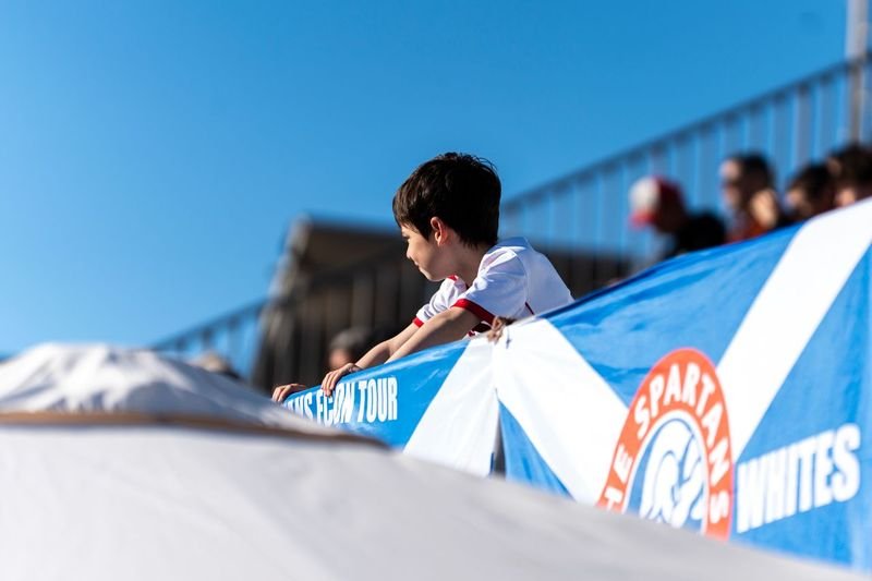 A young boy leaning on a railing at a sports event, with a blue and white tent and other spectators in the background, under a clear blue sky.