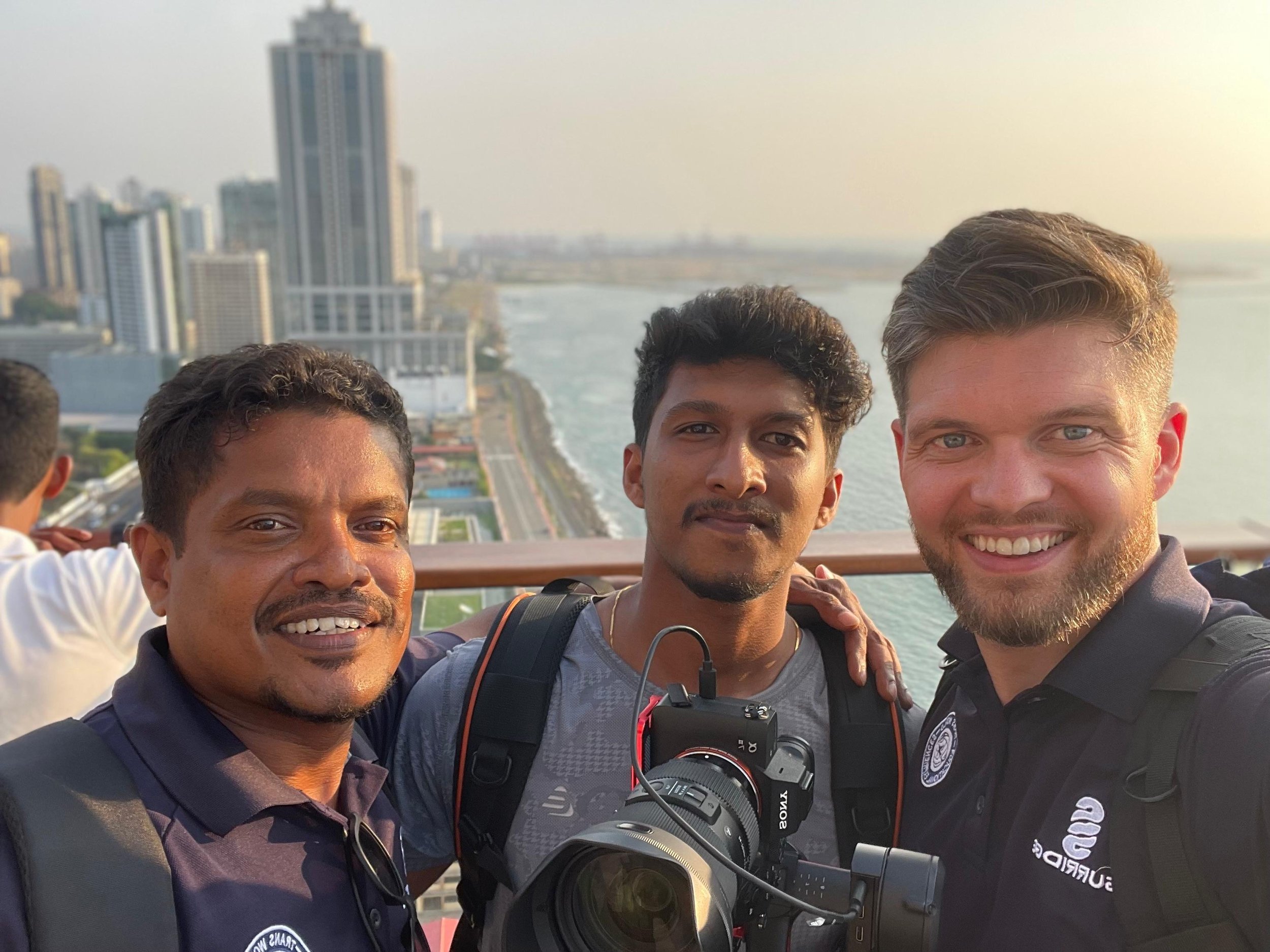 Four men standing close together taking a selfie, with a city skyline and river in the background during sunset.