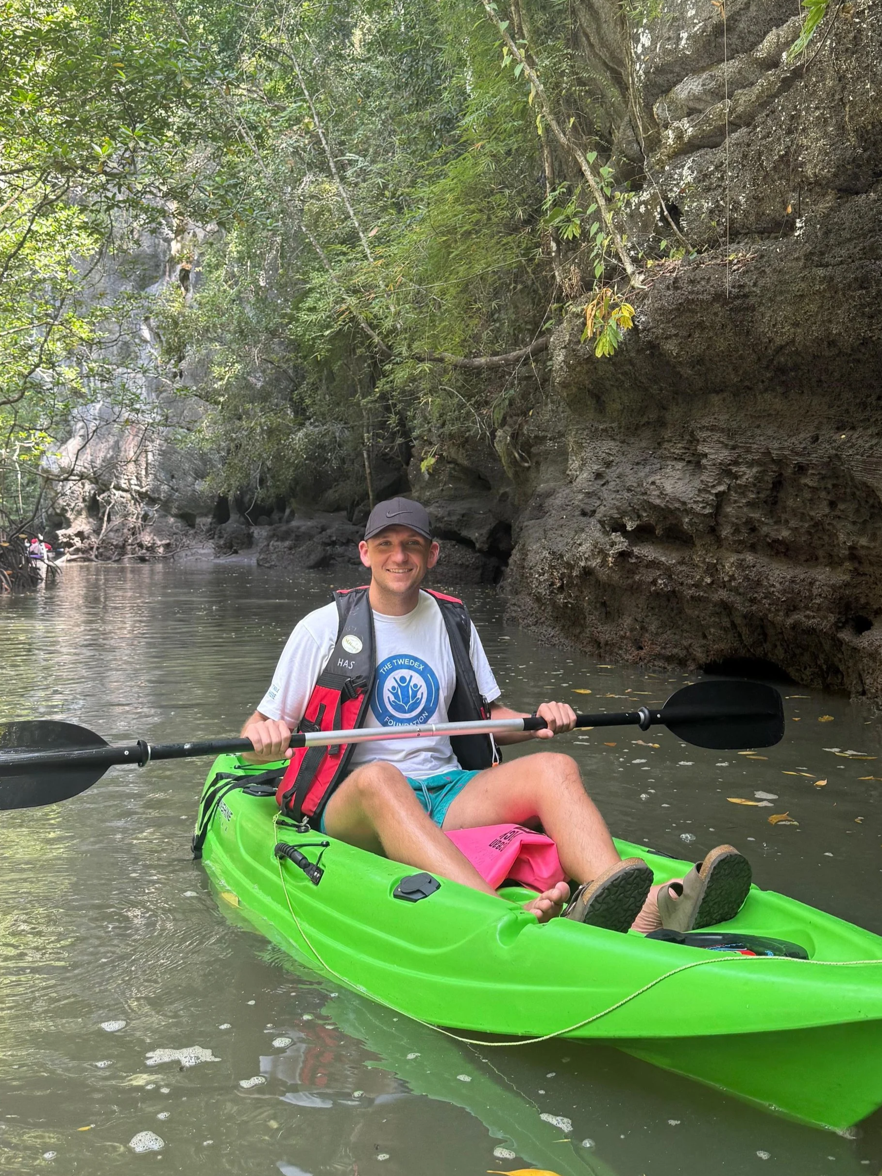 A man in a grey t-shirt, pink shorts, and a black cap, sitting in a bright green kayak on a calm river surrounded by lush green trees and large rocky cliffs.