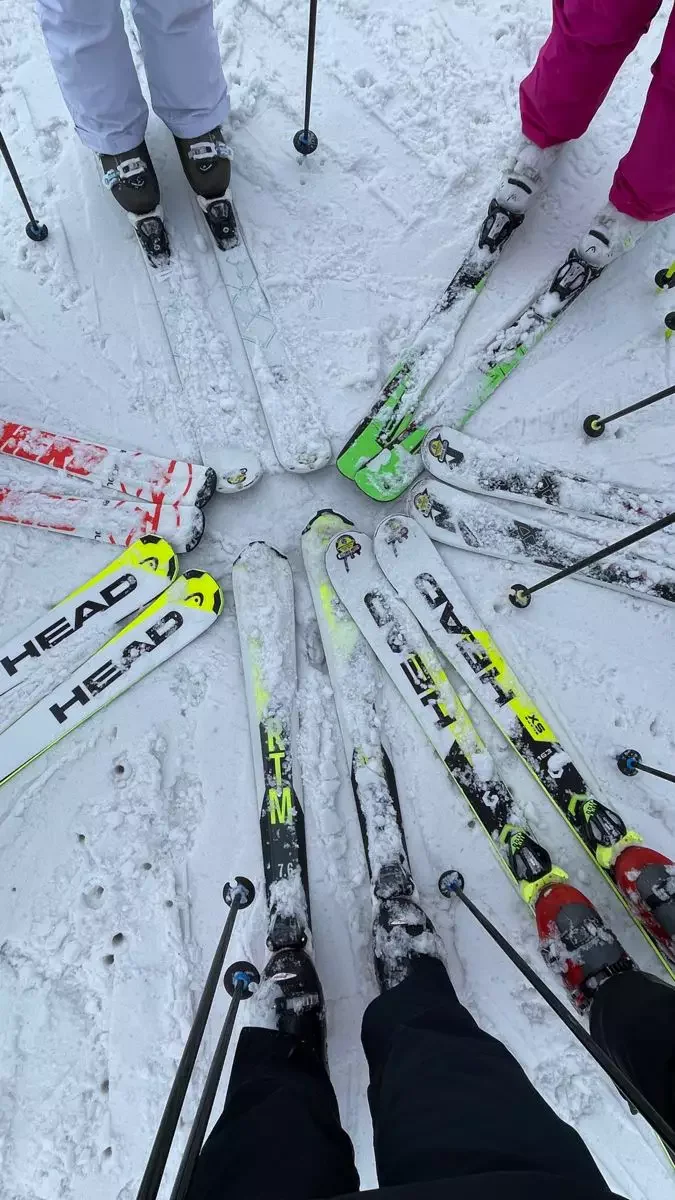 Group of people in ski boots, skis, and snow gear standing in a circle on snow-covered ground, preparing to ski.