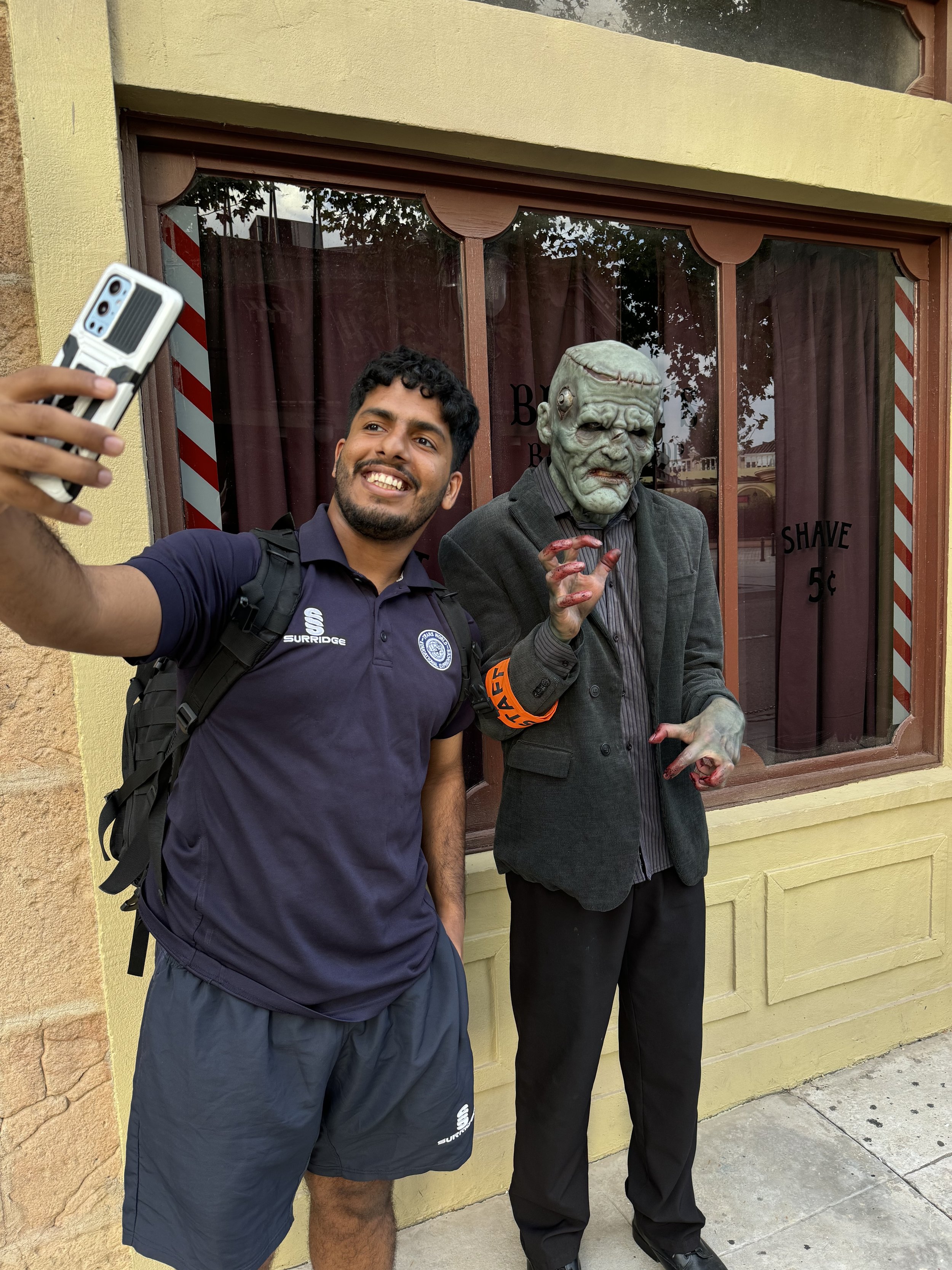 A man in a navy blue shirt and shorts taking a selfie with a person in a horror costume with a grotesque mask and bloody hands outside a building with yellow walls and a window.